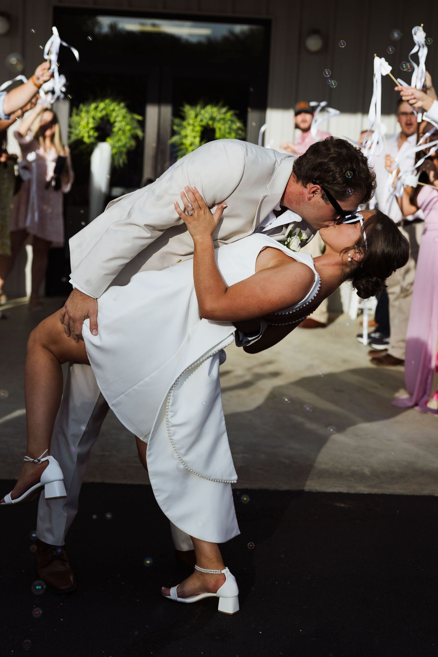 Groom dips bride in white dress for romantic kiss outside wedding venue surrounded by guests.