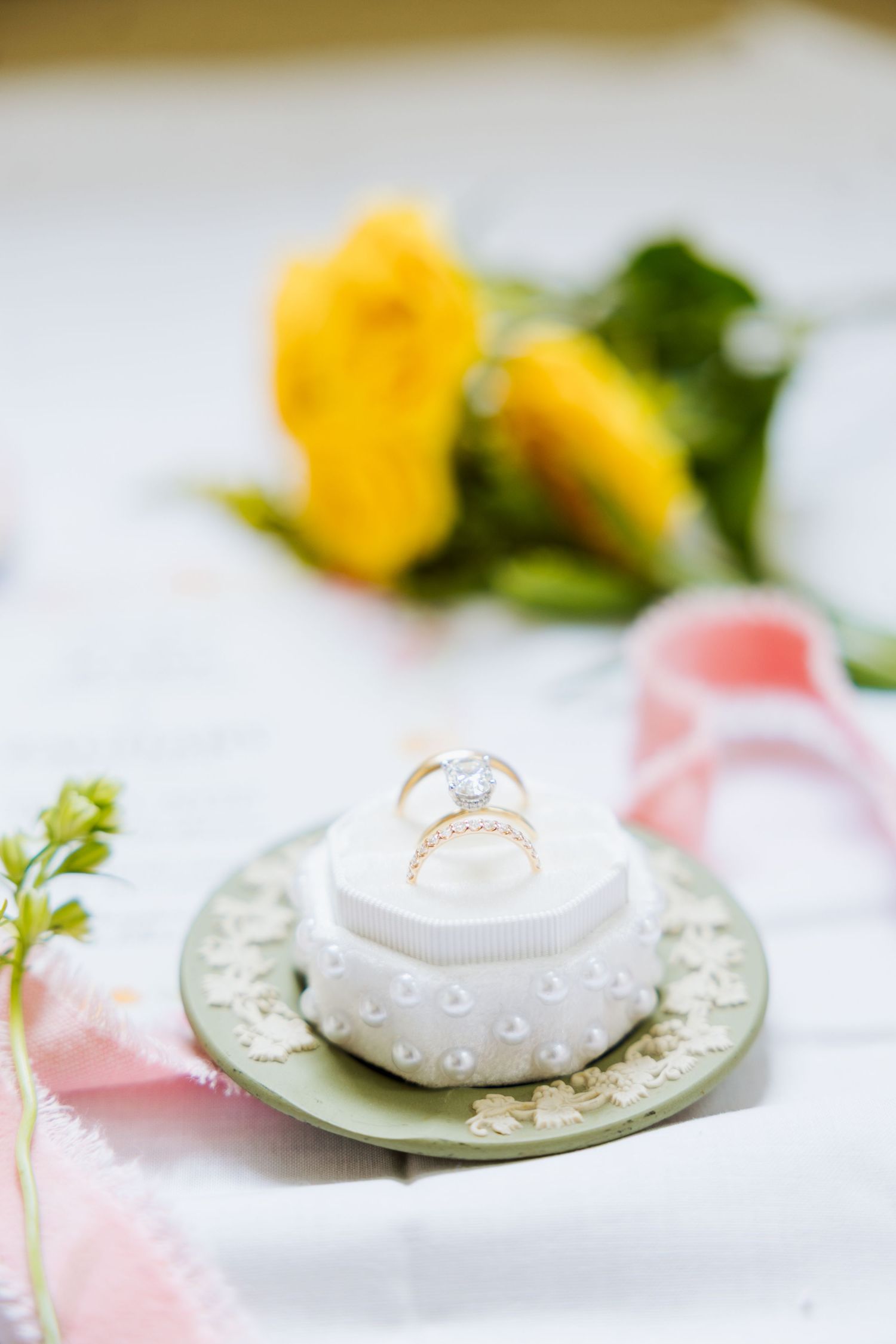 Gold diamond engagement ring displayed on white ceramic ring holder with green plate, pink ribbon and yellow roses in background.