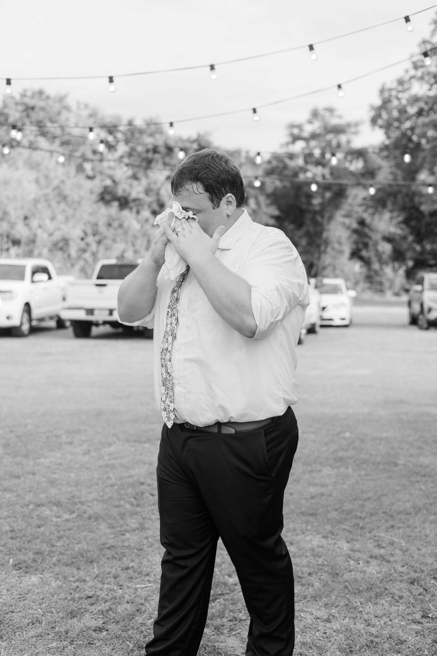 Emotional groom wiping tears with handkerchief outdoors at wedding reception with string lights.