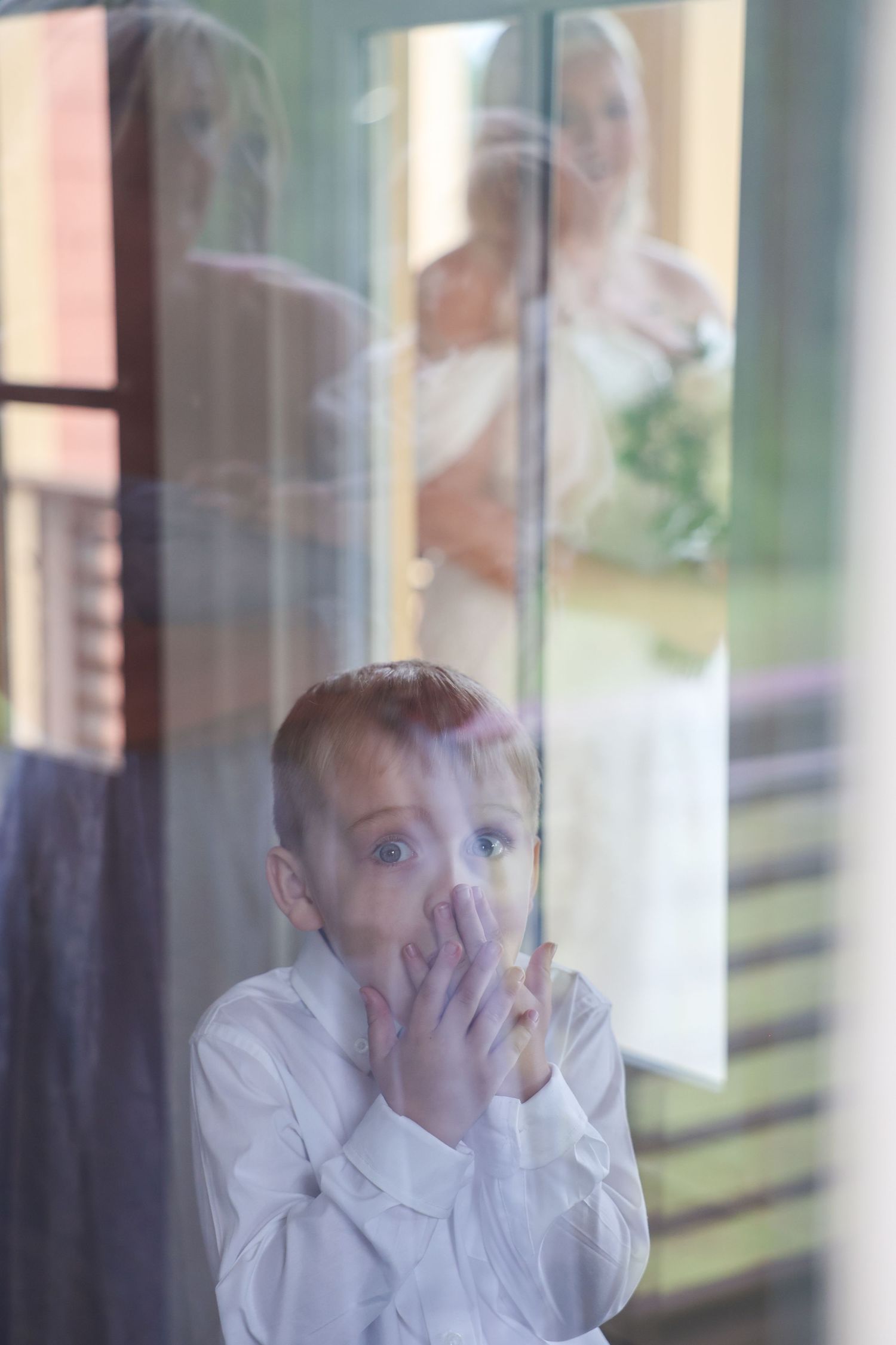 Surprised young boy in white shirt presses hands to face against window at wedding ceremony.