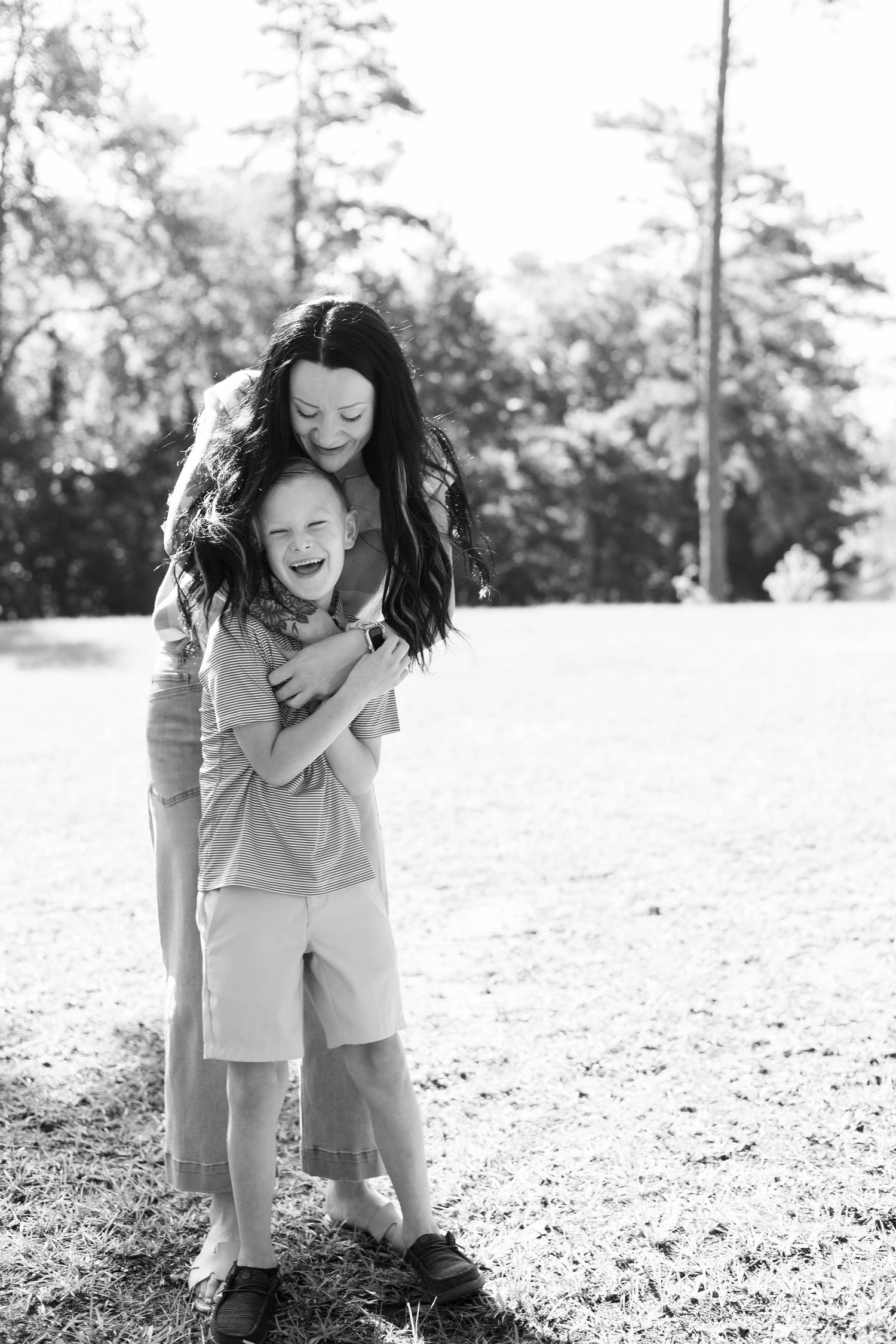 Black and white photo of a laughing boy being hugged from behind by a smiling woman in a sunny park.