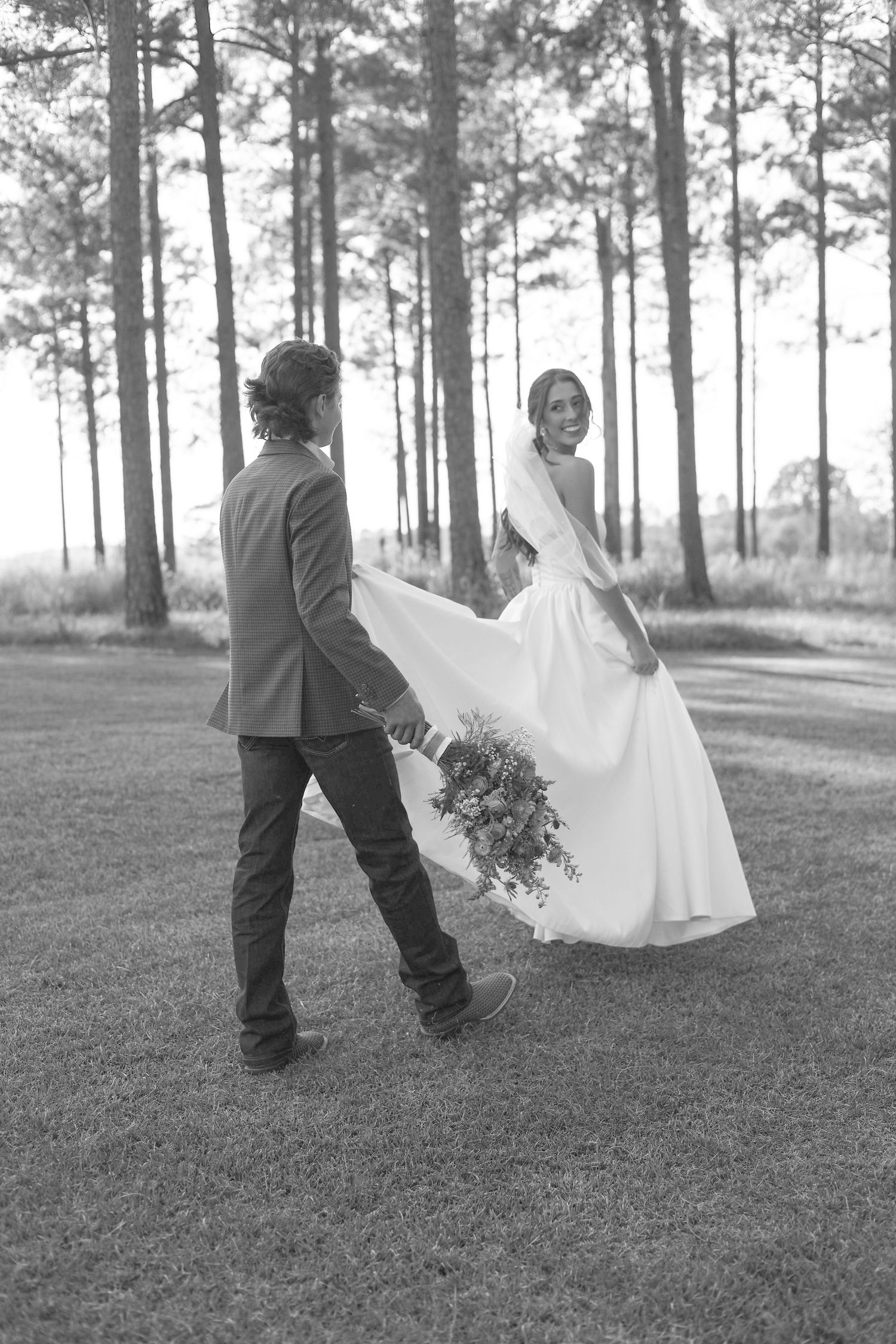 Black and white photo of a bride and groom walking hand in hand through a pine forest, bride holding a bouquet.