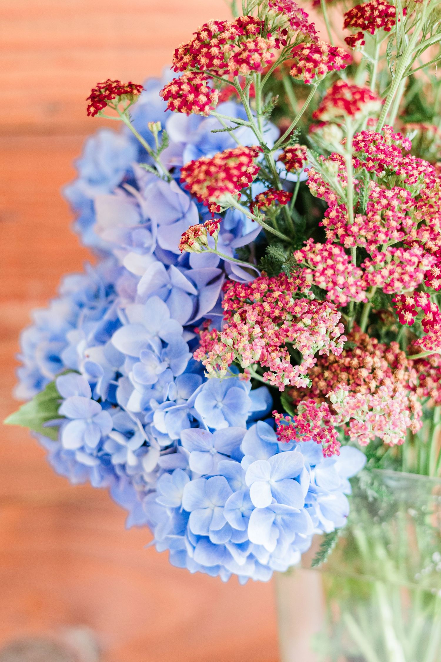 Blue hydrangea and red yarrow flowers arranged in a glass vase against a warm wooden background.