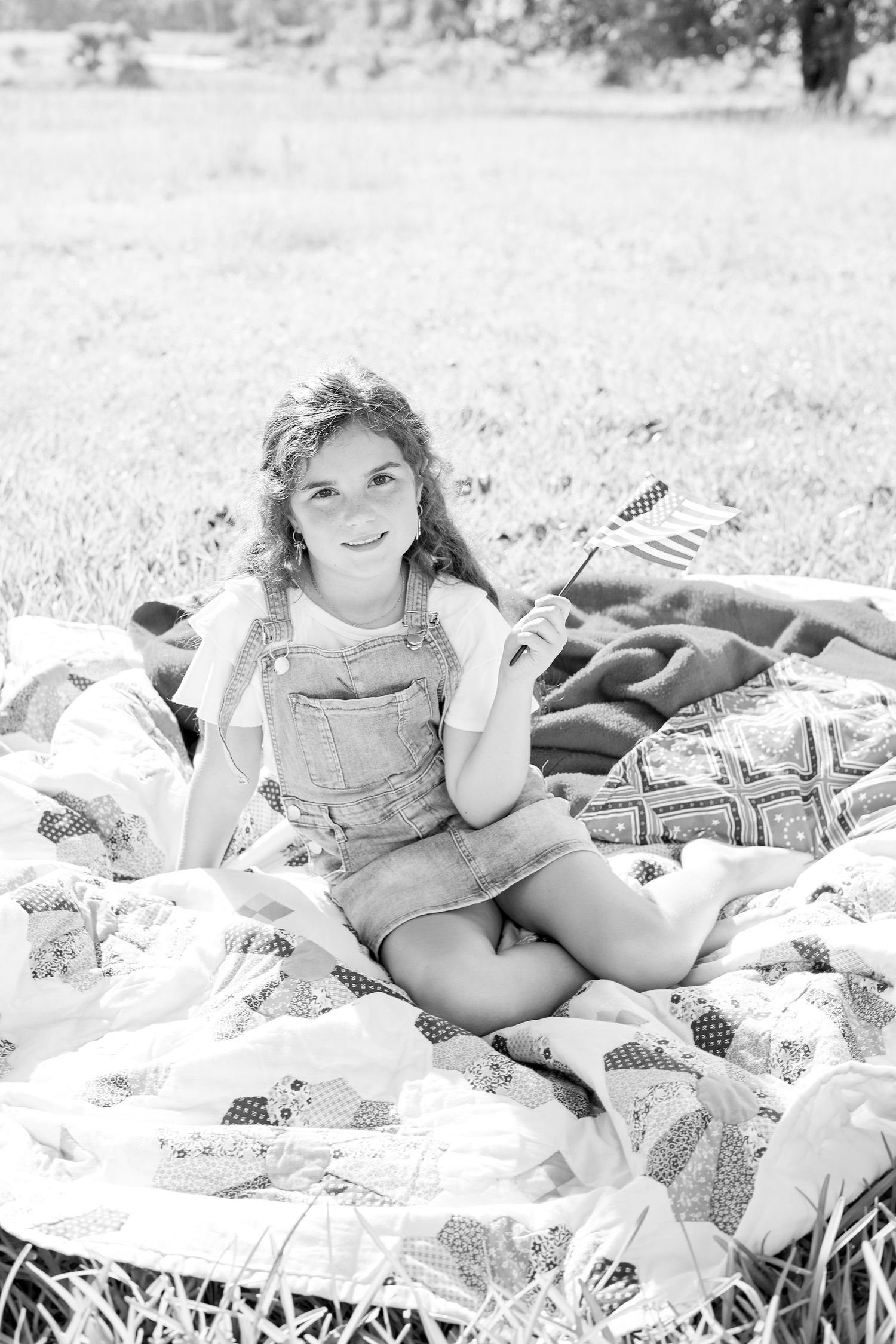Young girl sitting on a quilt blanket outdoors in a field, smiling and holding a small American flag.