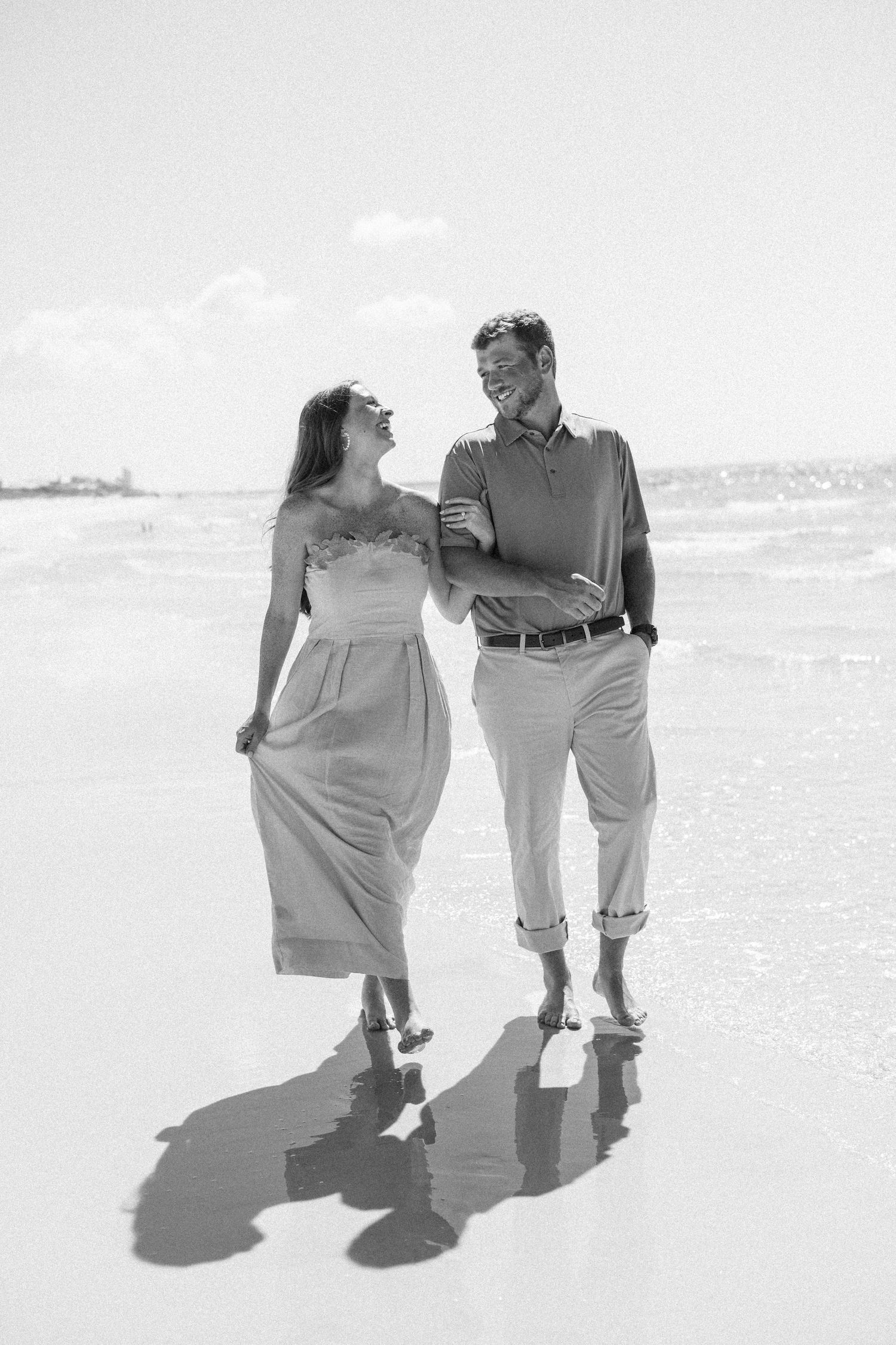 Happy couple walking hand in hand along a sunny beach shore in elegant engagement photos.