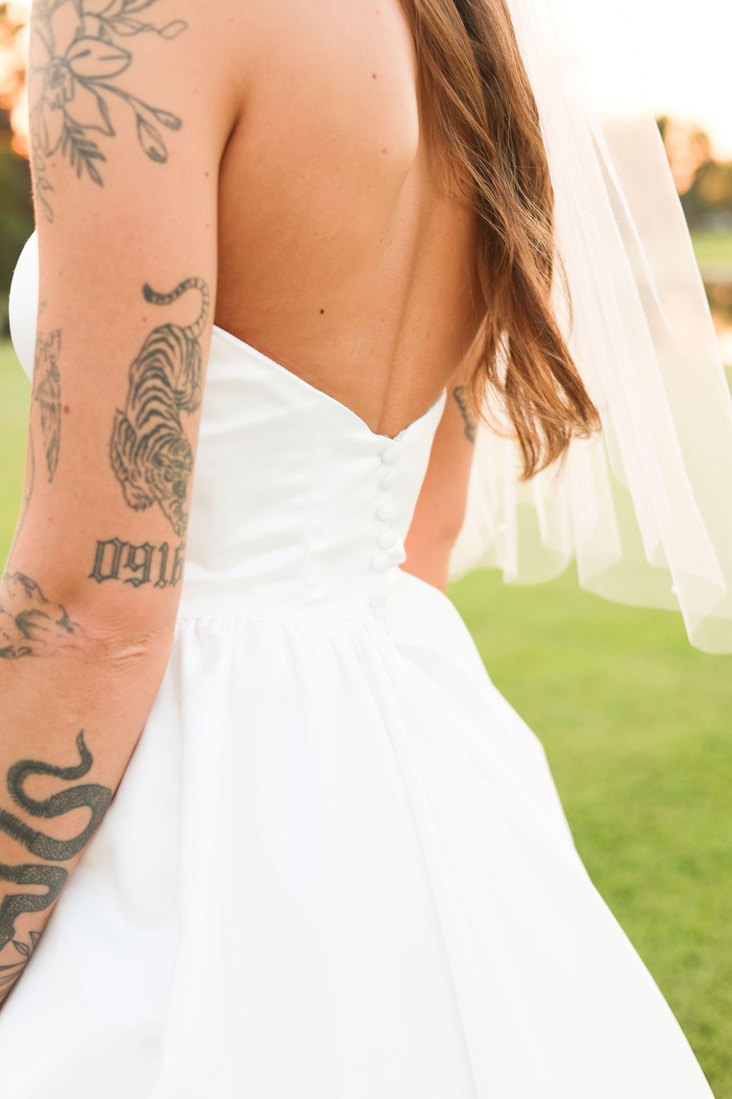 Elegant white wedding dress with visible arm tattoos shown in detail against a sunset backdrop.