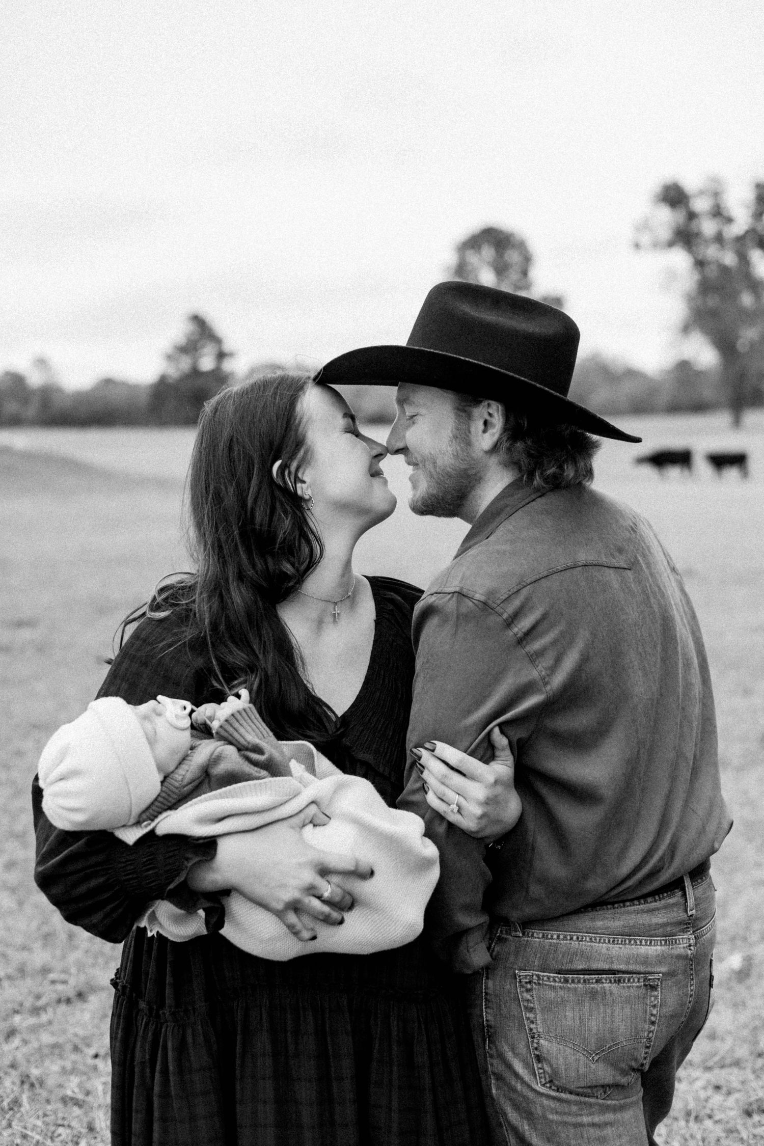 Parents embrace while holding their newborn baby in a pastoral field setting.