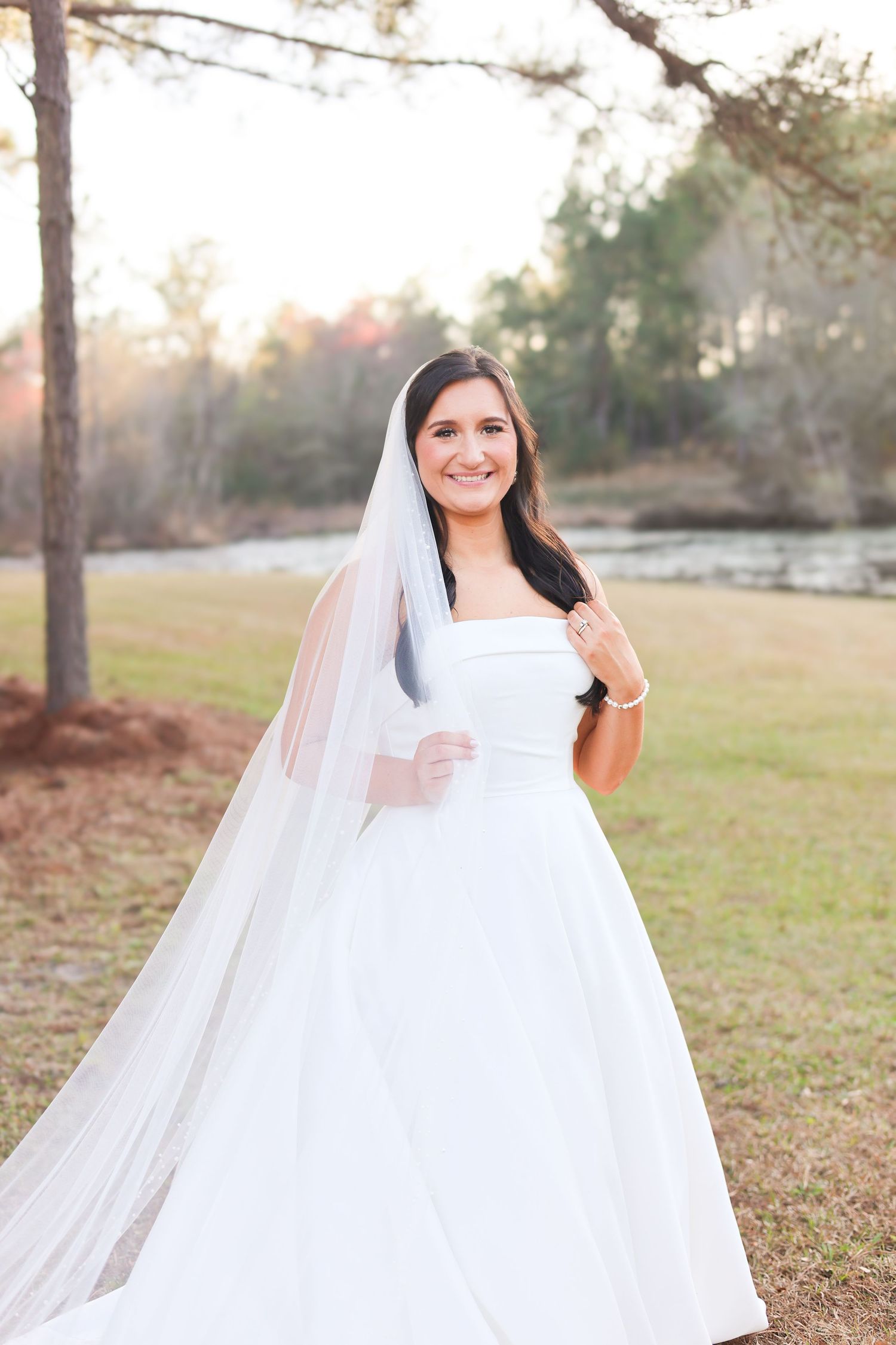 Smiling bride in strapless white ball gown and cathedral veil posing outdoors among pine trees at golden hour.