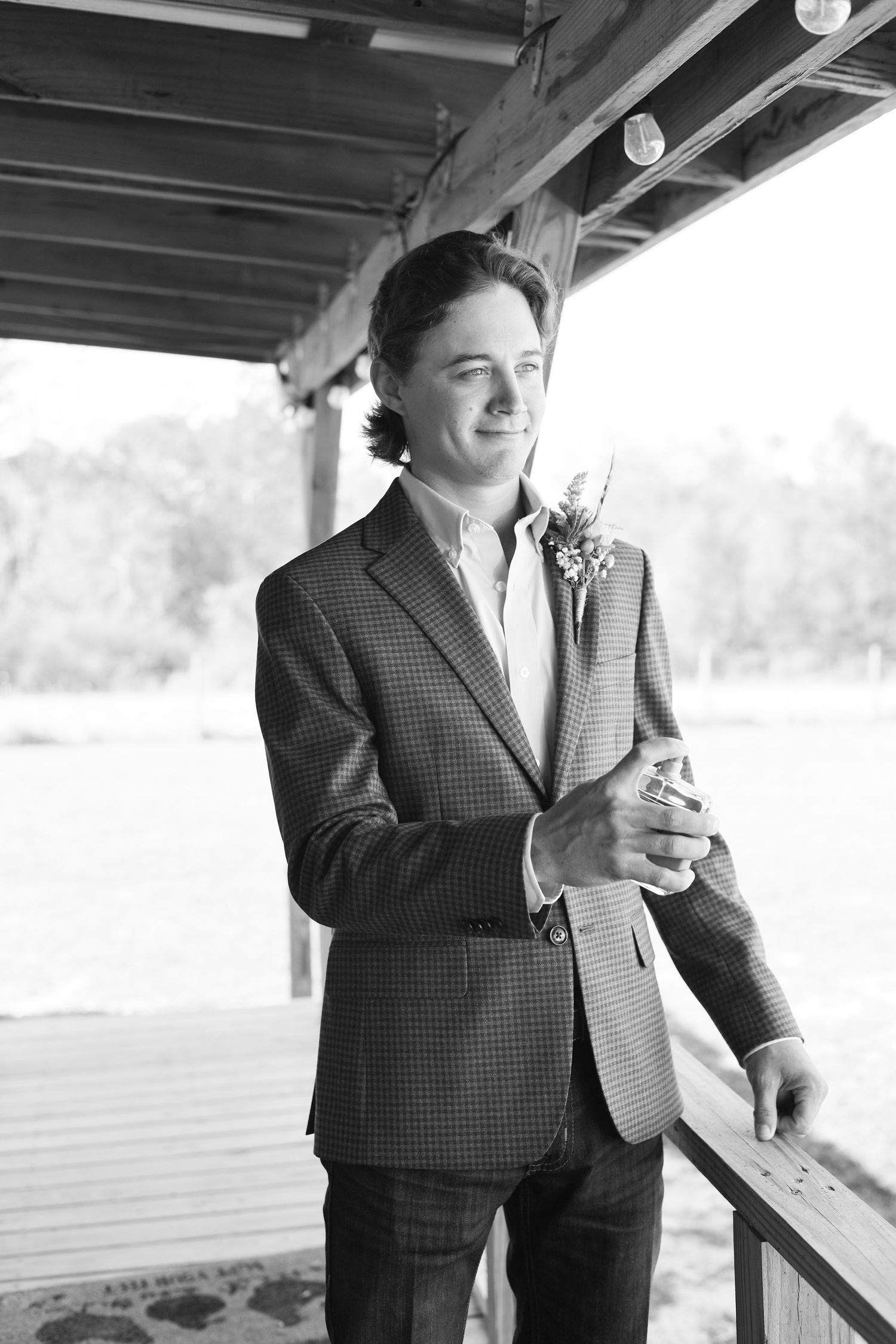 A young man in a plaid blazer with a boutonniere smiles while standing on a rustic wooden porch.