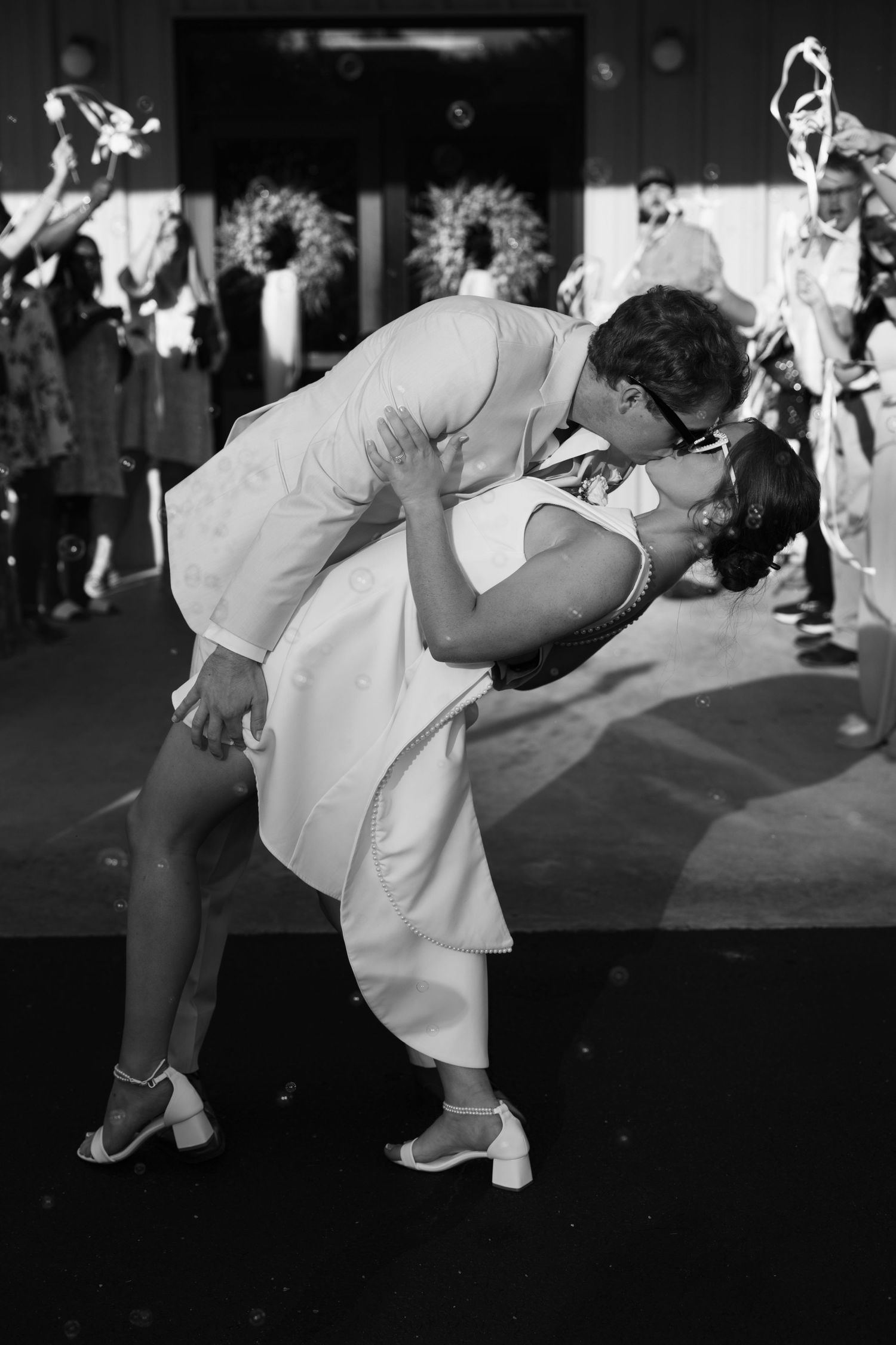 Black and white photo of a couple sharing a romantic dip kiss at a wedding reception with guests celebrating behind them.