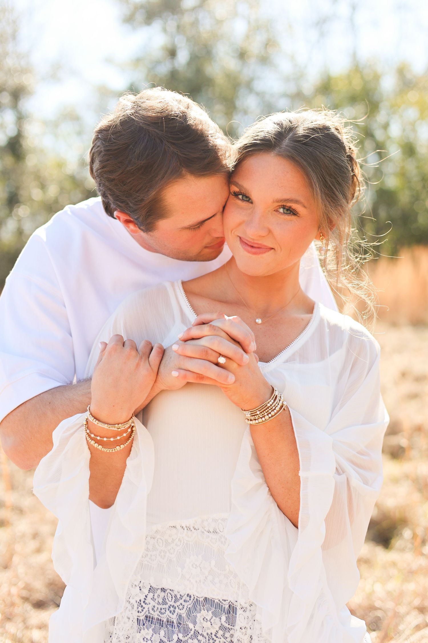 A romantic couple in white outfits share tender kisses in a sunlit outdoor setting with a dreamy golden glow.