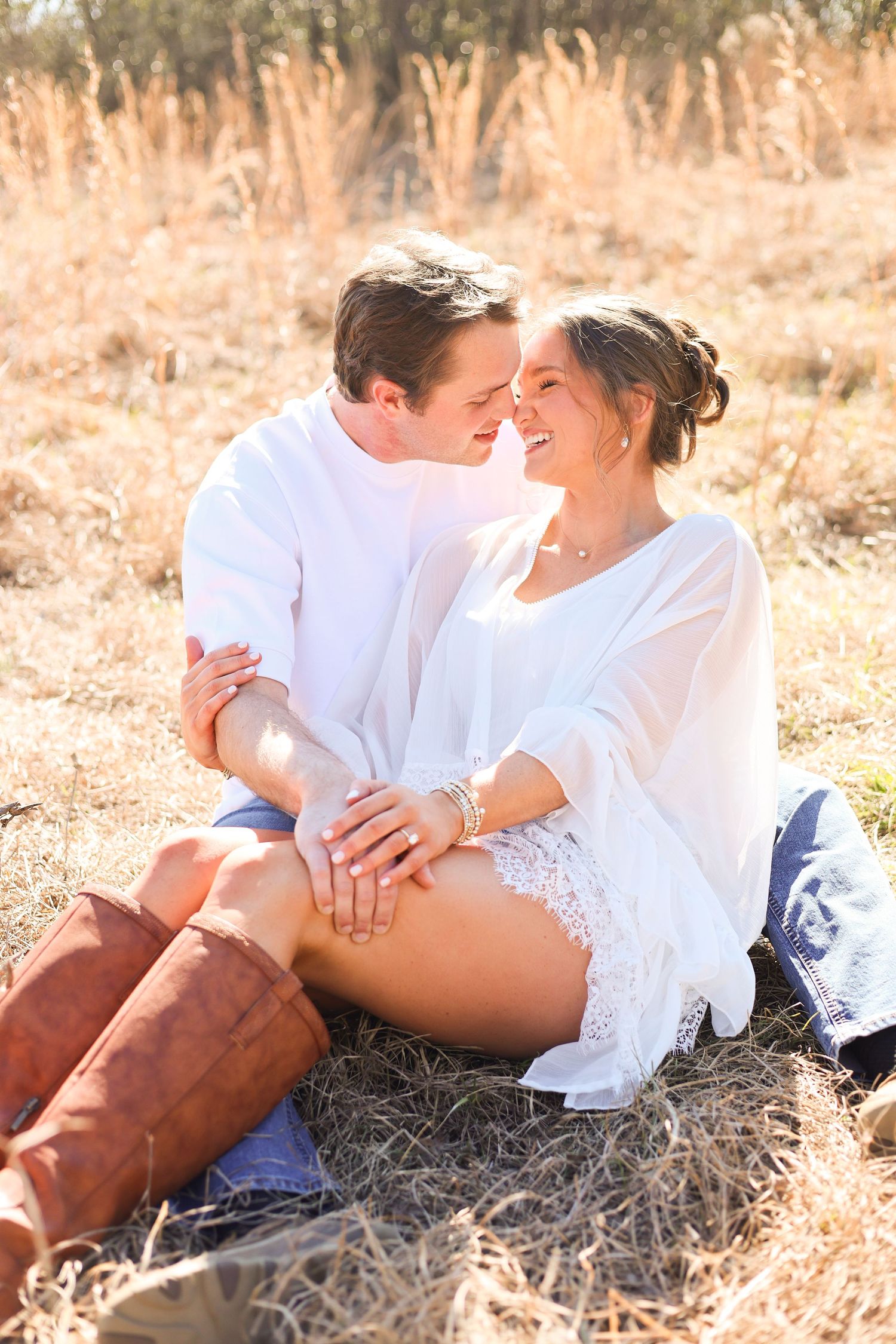 Romantic couple in white clothing share an intimate moment while sitting in a sunlit field with tall golden grass.