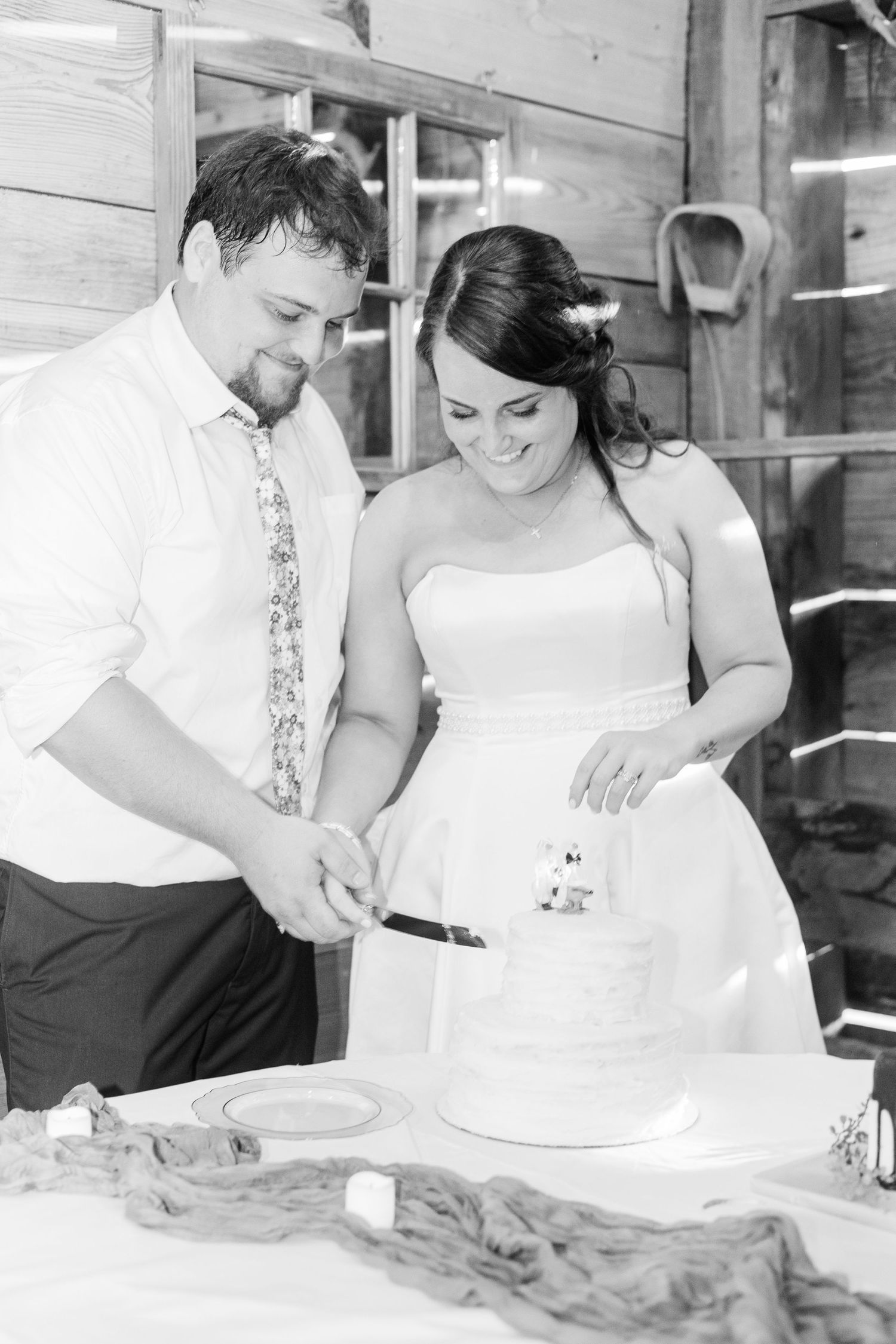 Black and white photo of bride and groom cutting wedding cake at rustic barn reception.