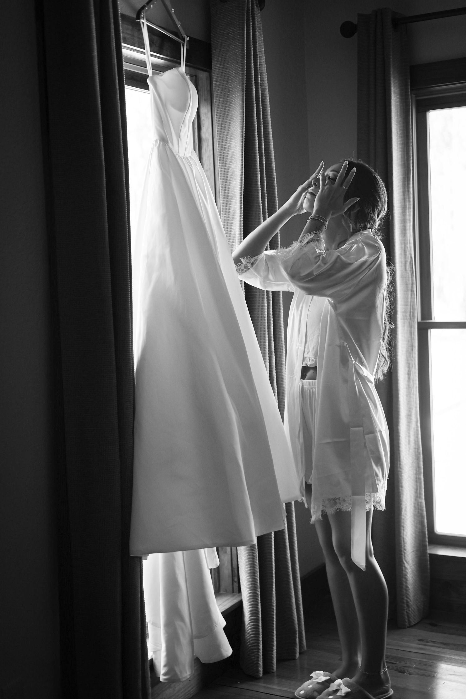 Bride in white robe admiring her wedding gown hanging by window in black and white photo.