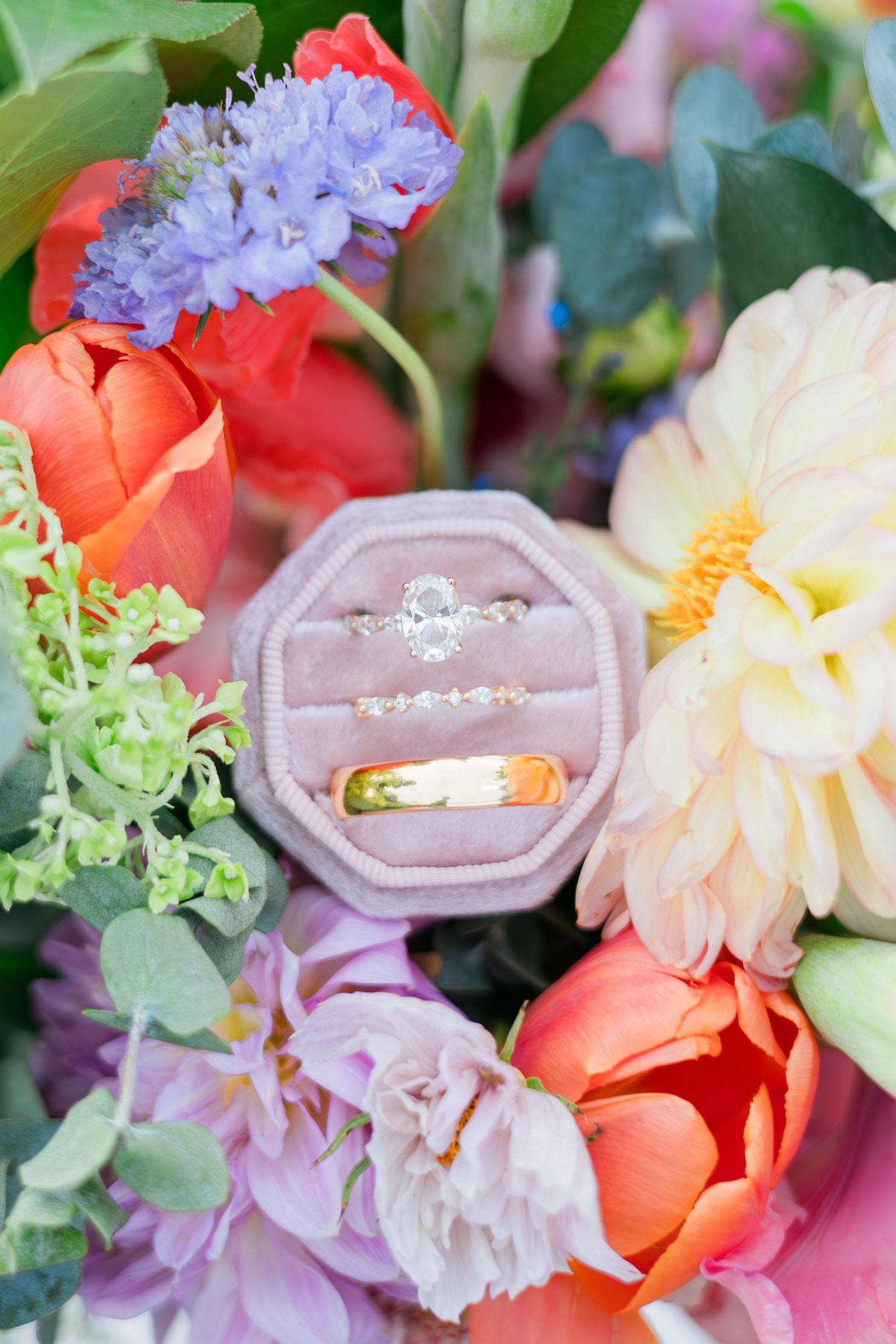 Oval diamond engagement ring and gold wedding band in pink velvet box surrounded by colorful spring flowers.
