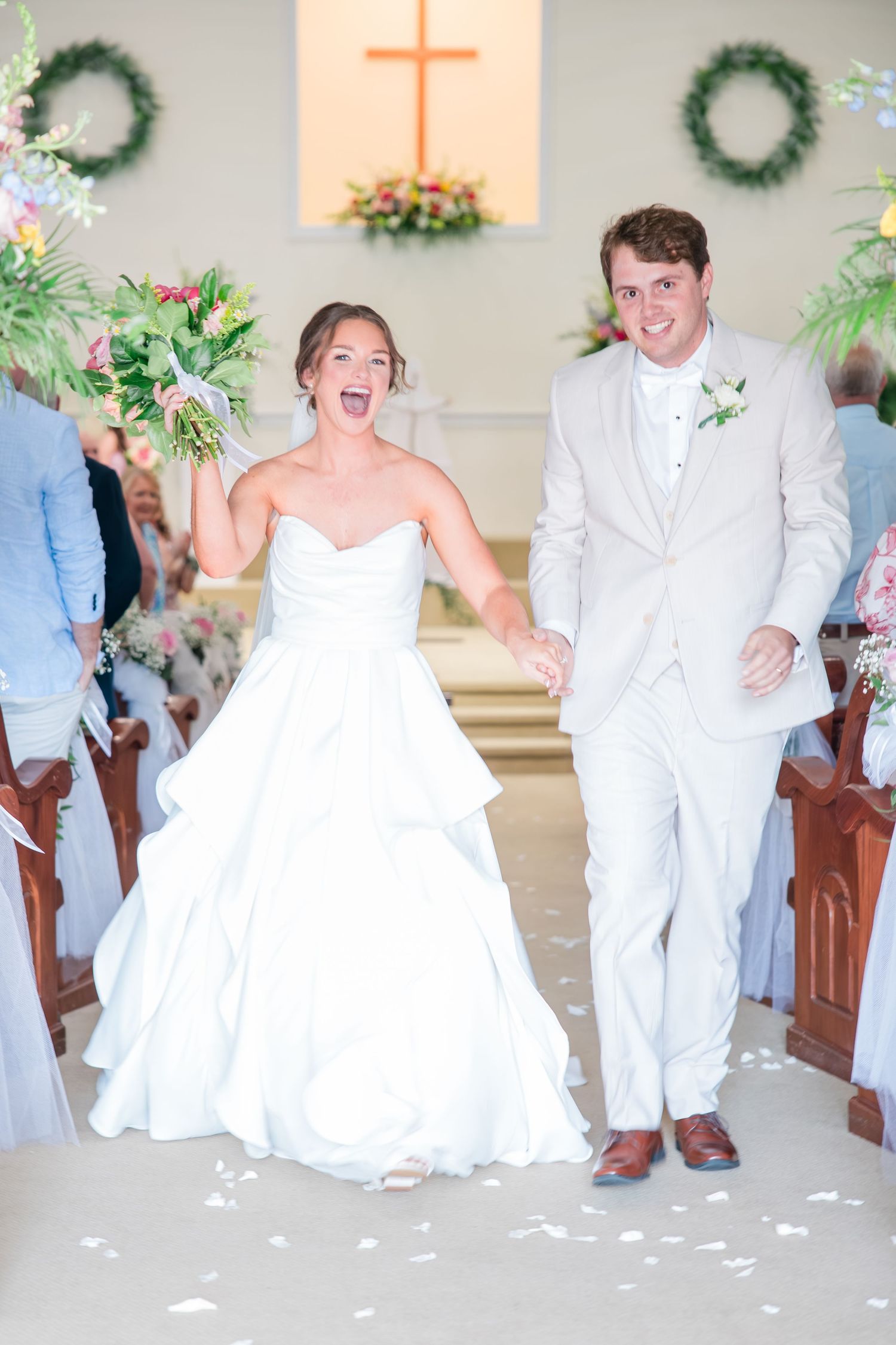 Joyful bride and groom walk down church aisle after wedding ceremony, bride in white gown raising bouquet.