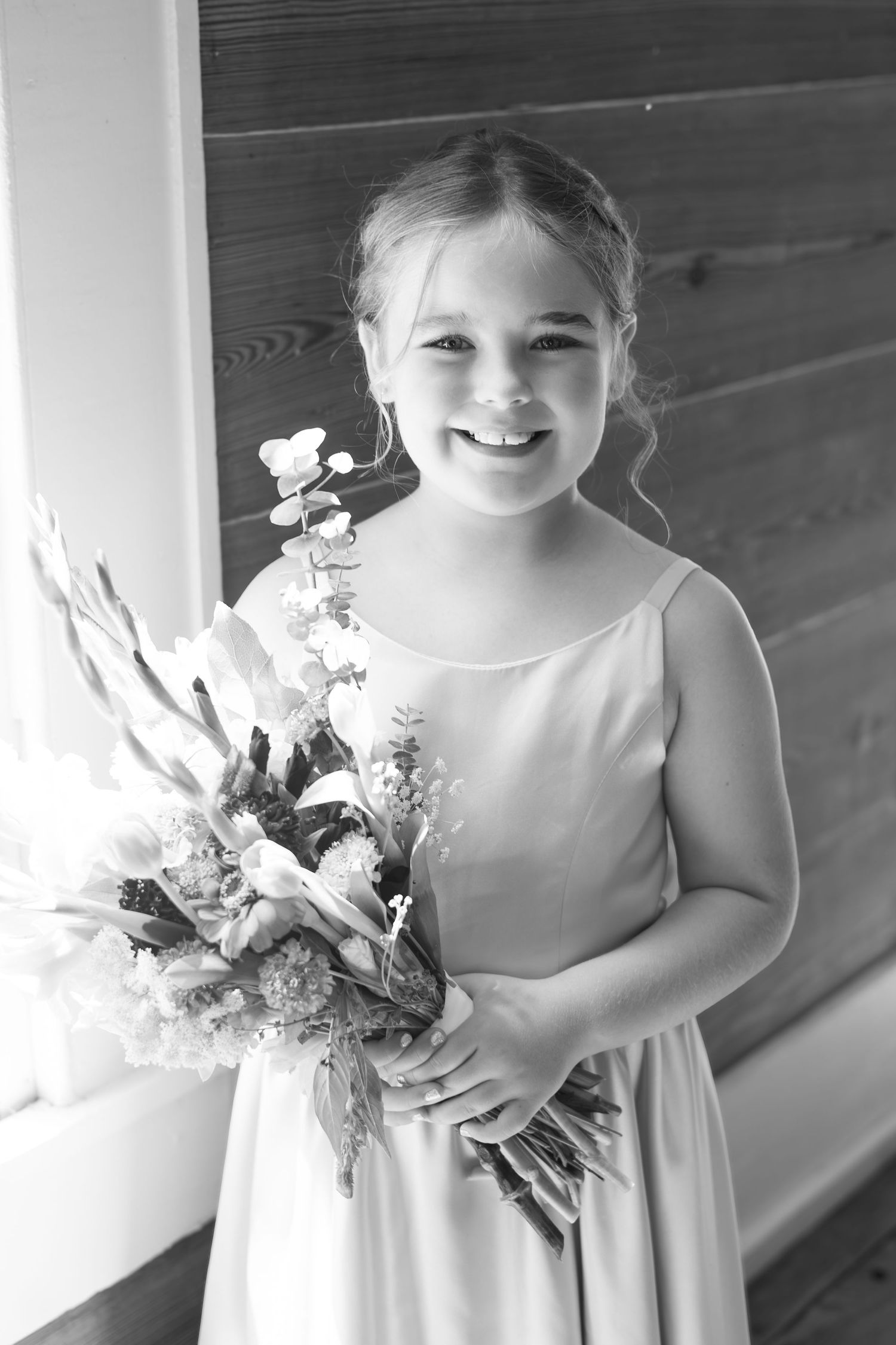 Smiling flower girl in white dress holding a bouquet at a wedding ceremony, black and white photo.
