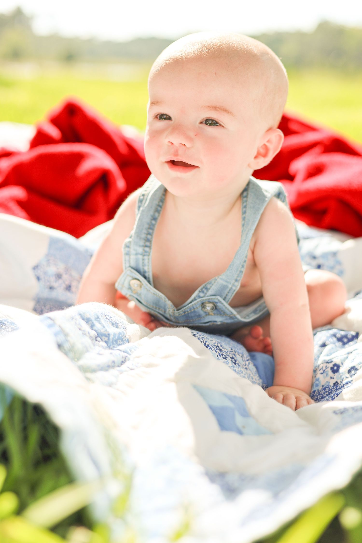 Adorable baby in denim overalls sitting on a blue and white quilt outdoors, smiling with red blanket behind.