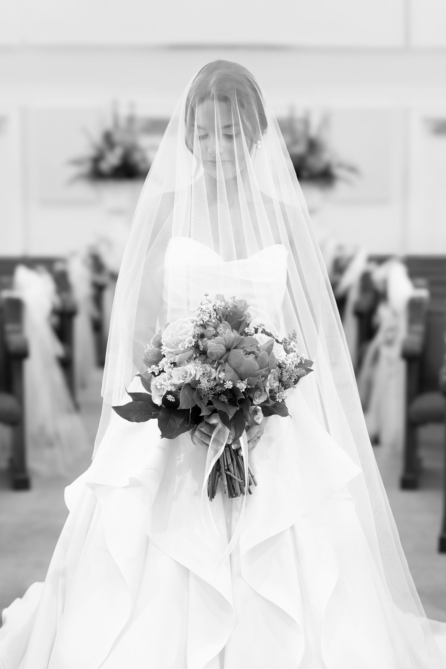Elegant bride in white gown and veil holding floral bouquet standing in church aisle black white.