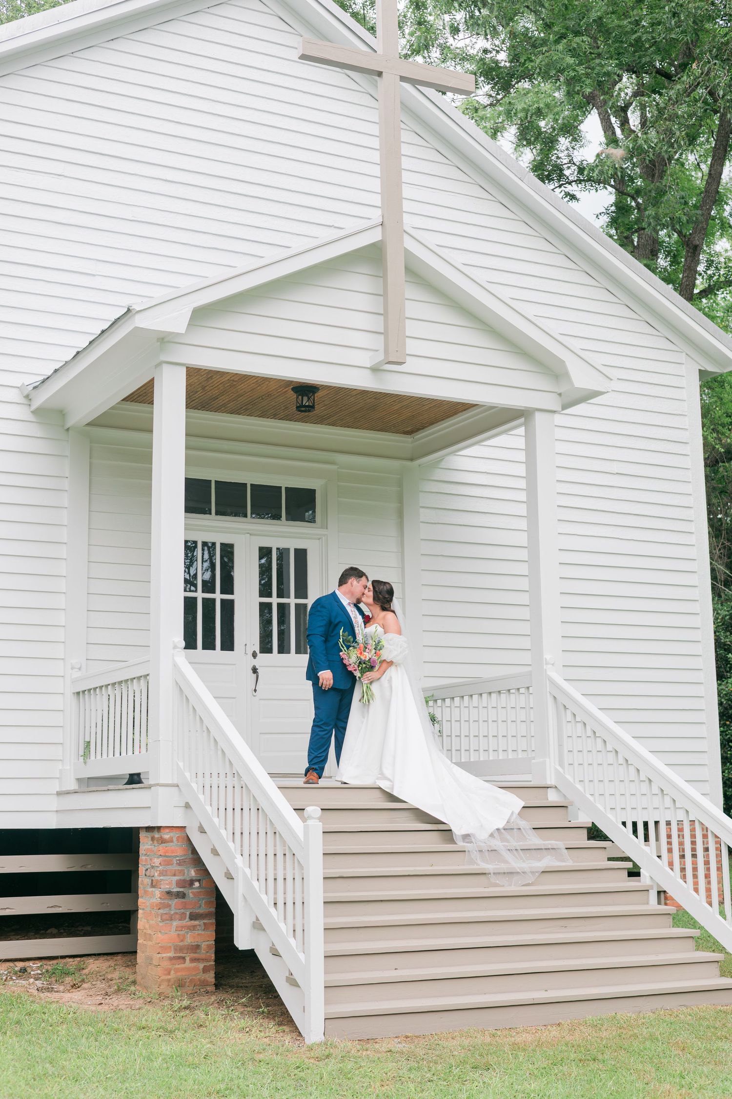 Bride and groom kissing on steps of white chapel with cross, bride in flowing white dress holding colorful bouquet.