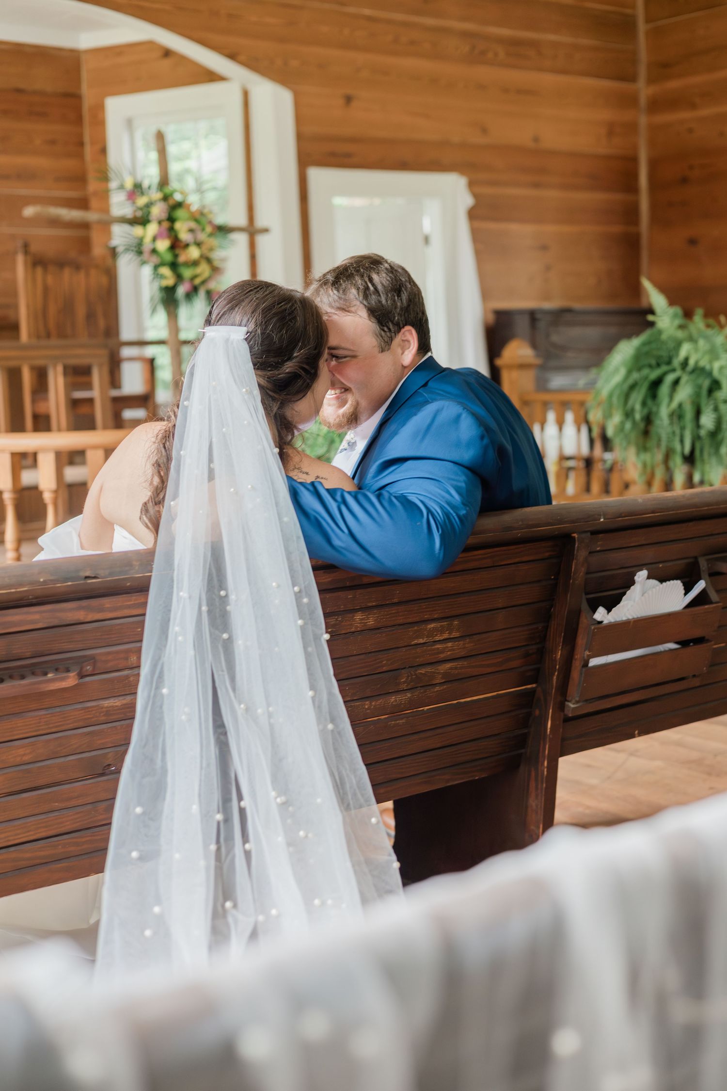 Bride and groom share an intimate moment on a wooden church pew, bride wearing pearl-adorned veil in rustic chapel.