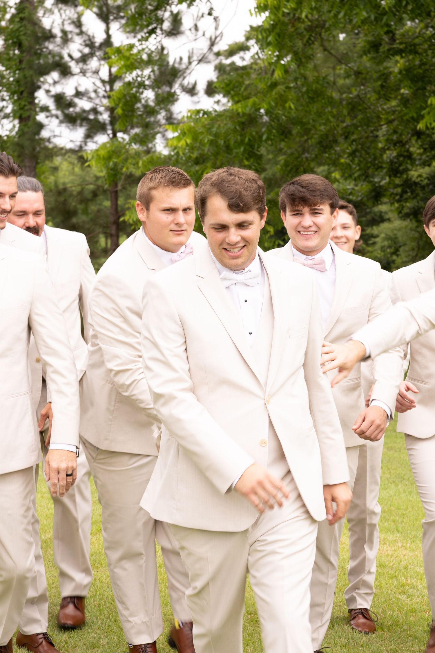 Groom and groomsmen in white suits with pink bow ties walking outdoors at a wedding celebration.