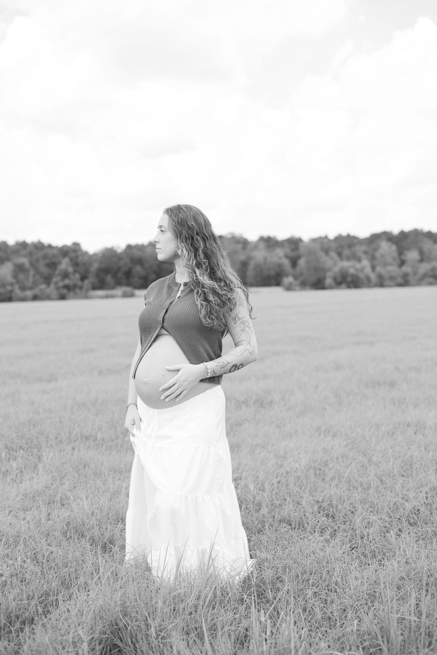 Black and white photo of a pregnant woman in a white skirt standing in a field, cradling her baby bump.