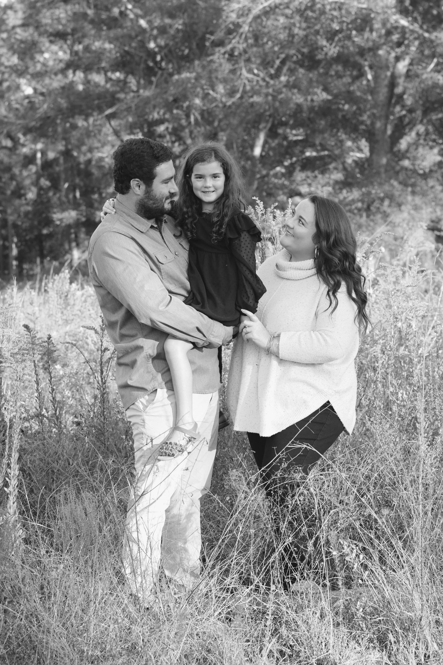 Black and white family portrait of parents and young daughter standing in tall grass with trees in background.