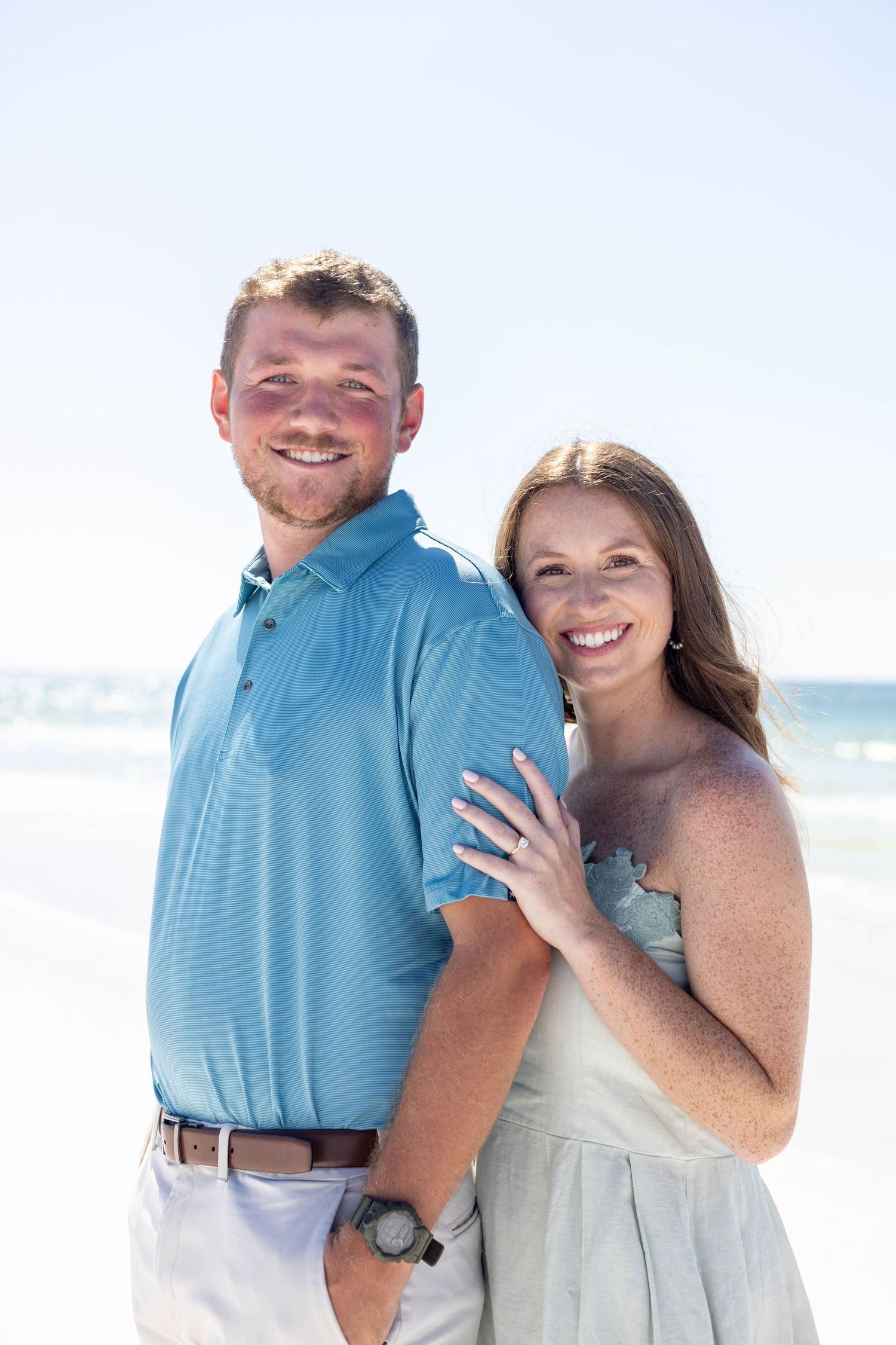 Happy couple smiling together on a sunny beach, woman in blue dress with hand on mans shoulder.