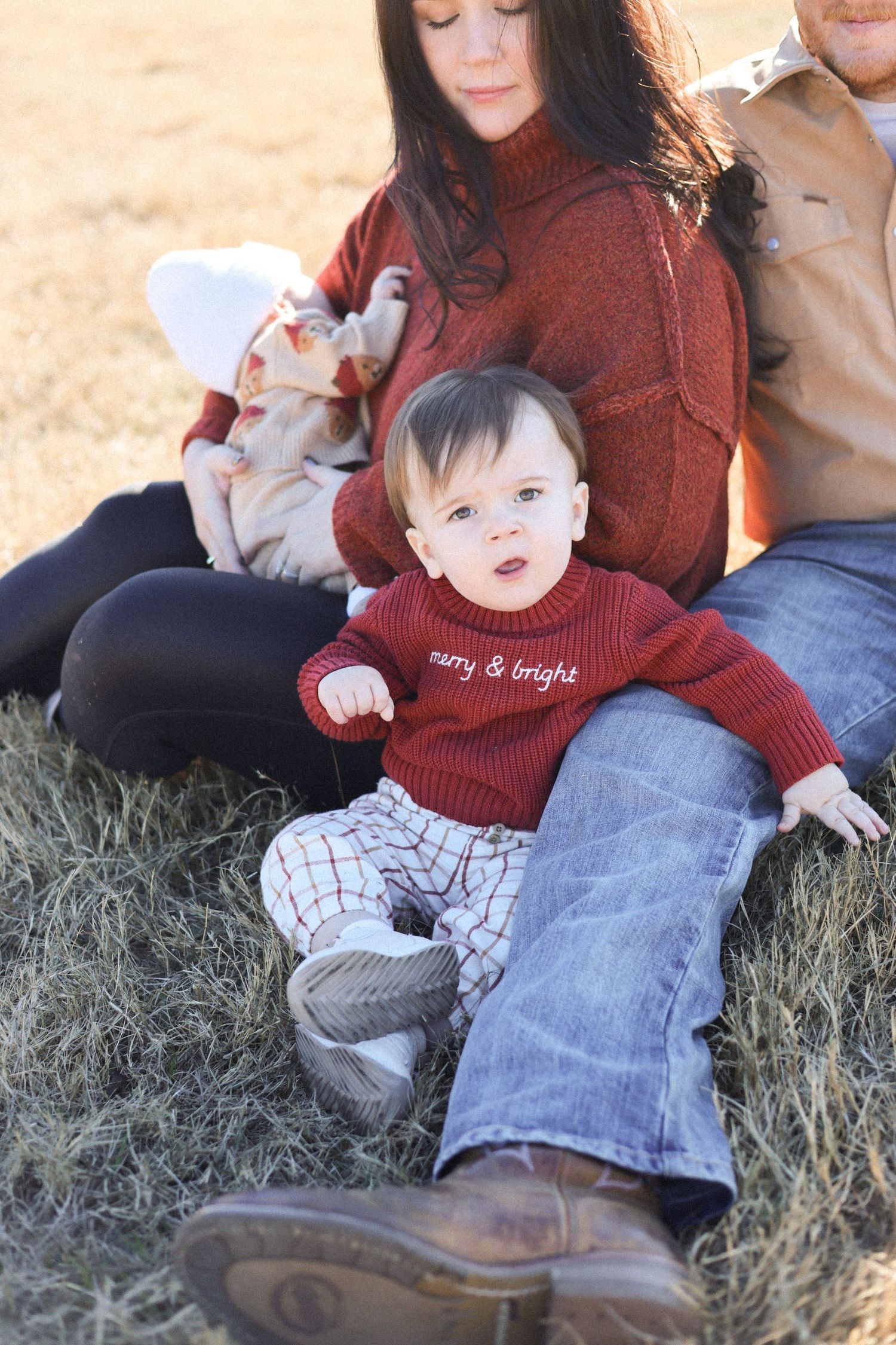 A family sits together on dried grass, with a baby wearing a red sweater and white patterned pants.