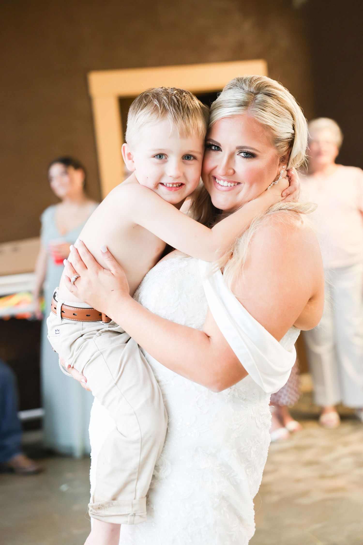 Smiling bride in white strapless gown hugs a young blond boy at wedding reception celebration.