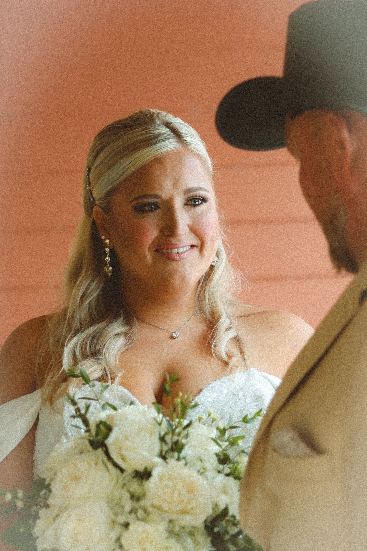 Radiant blonde bride smiling at groom holding white rose bouquet at outdoor wedding ceremony.