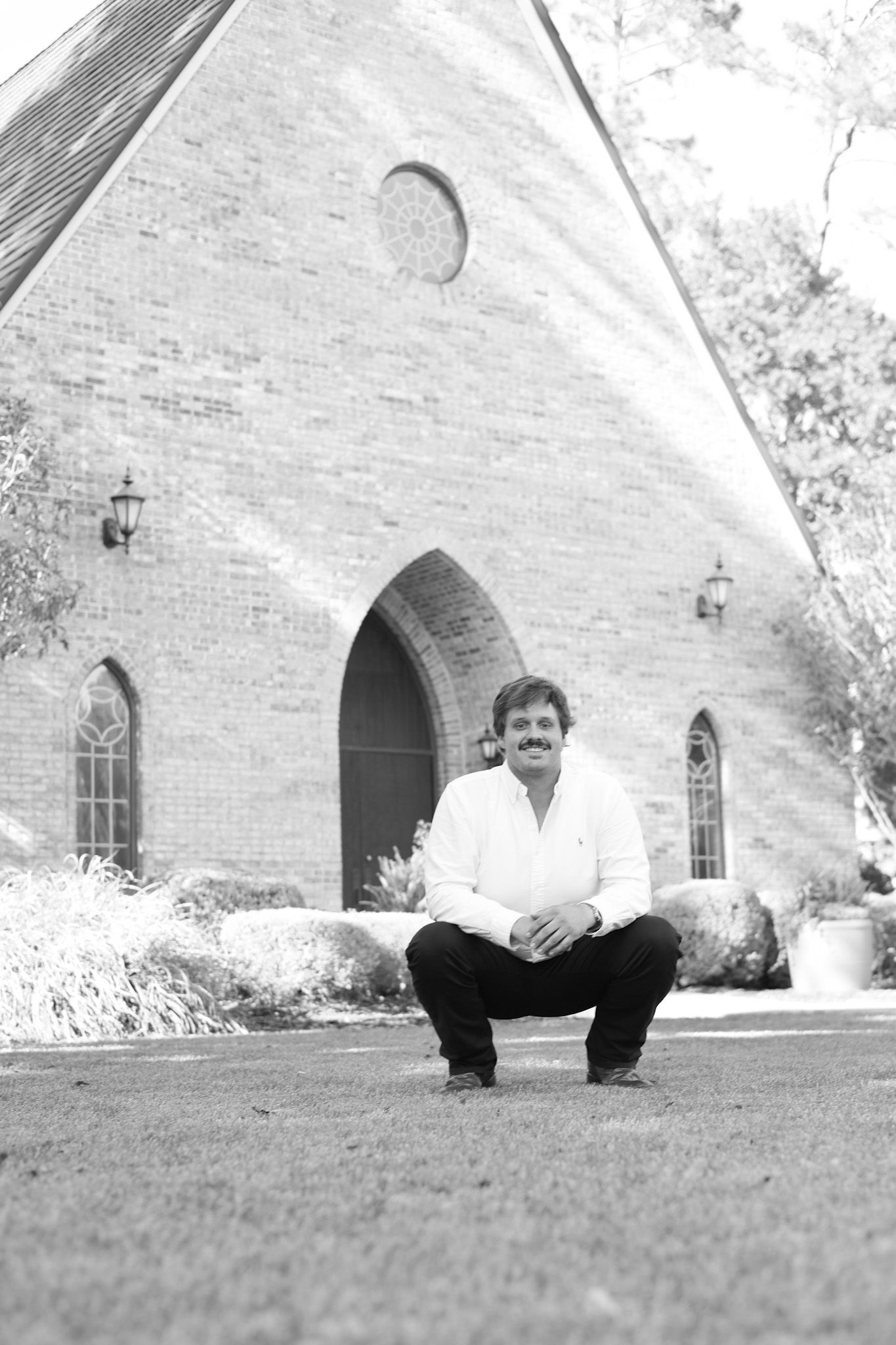 Black and white portrait of graduate sitting on grass near white church building.