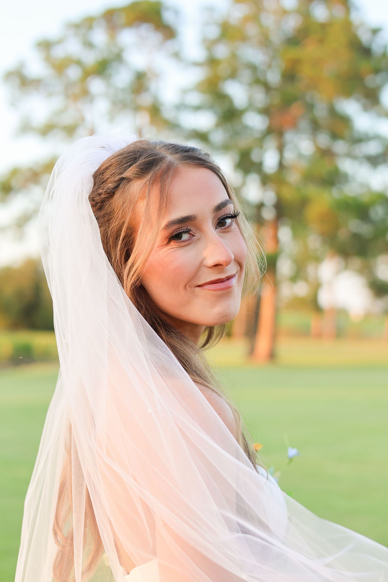 Ethereal bridal portrait with delicate veil captured in golden hour light on grassy field.