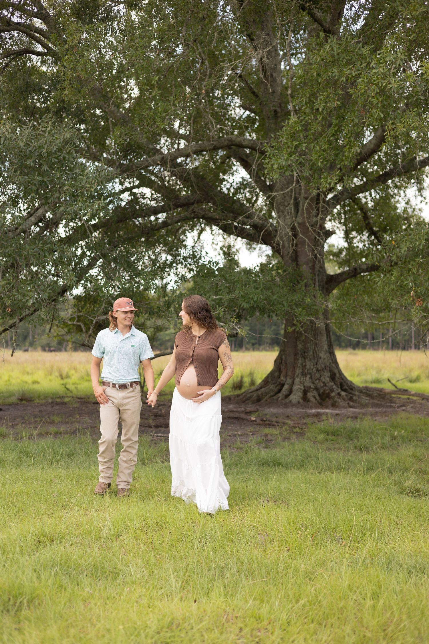Pregnant couple holding hands in a green meadow before a large oak tree during an outdoor maternity photo session.
