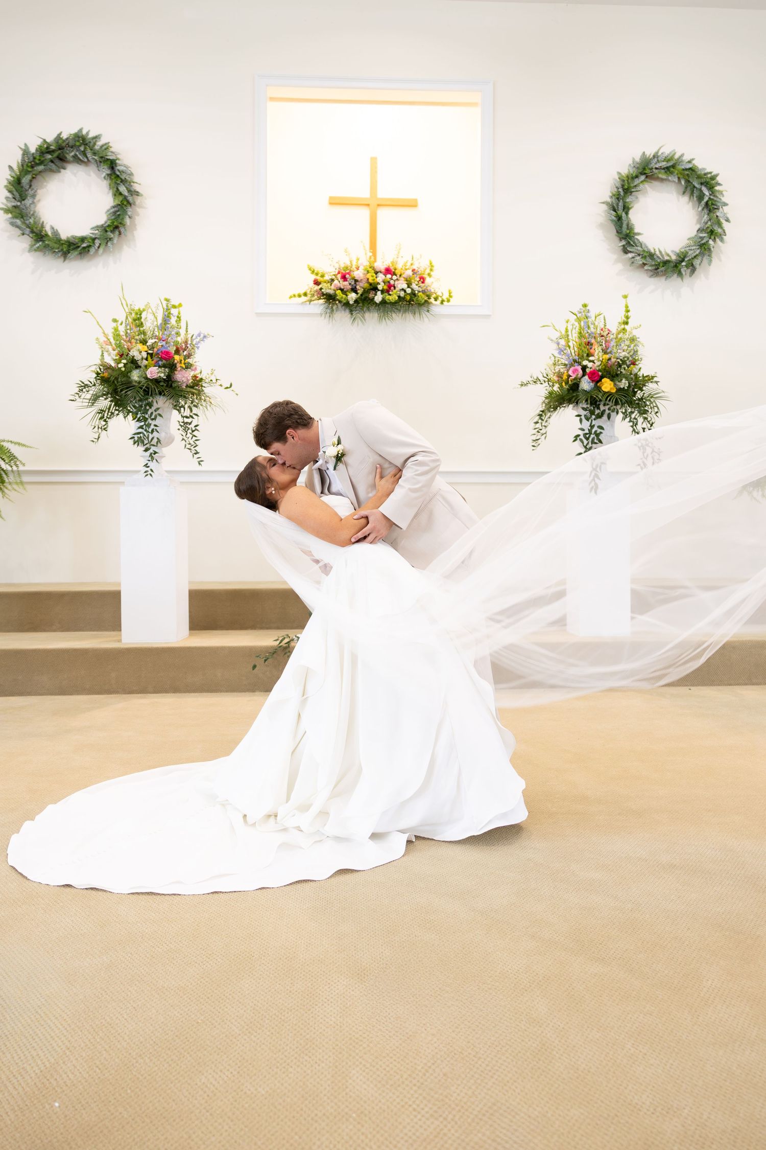 Bride and groom share romantic dip kiss at church altar with floral arrangements and cross backdrop.