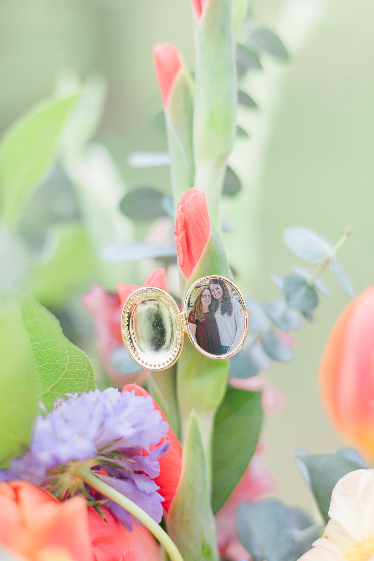 Open gold locket with couple photo charm nestled among vibrant coral tulips and purple flowers in bridal bouquet.