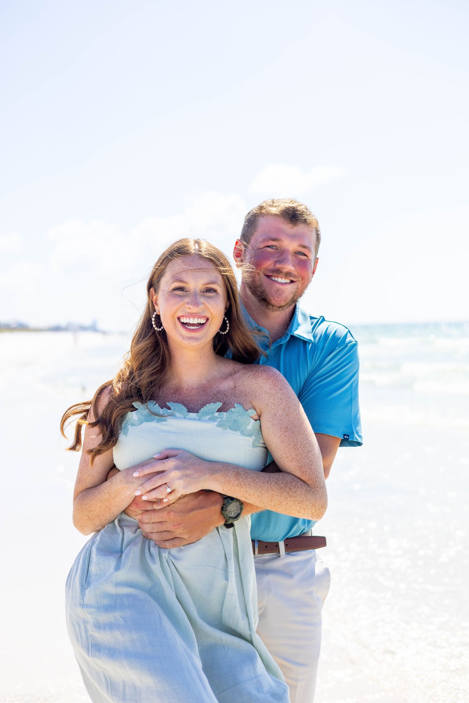 A happy couple embraces on a sunny beach with waves visible in the background.