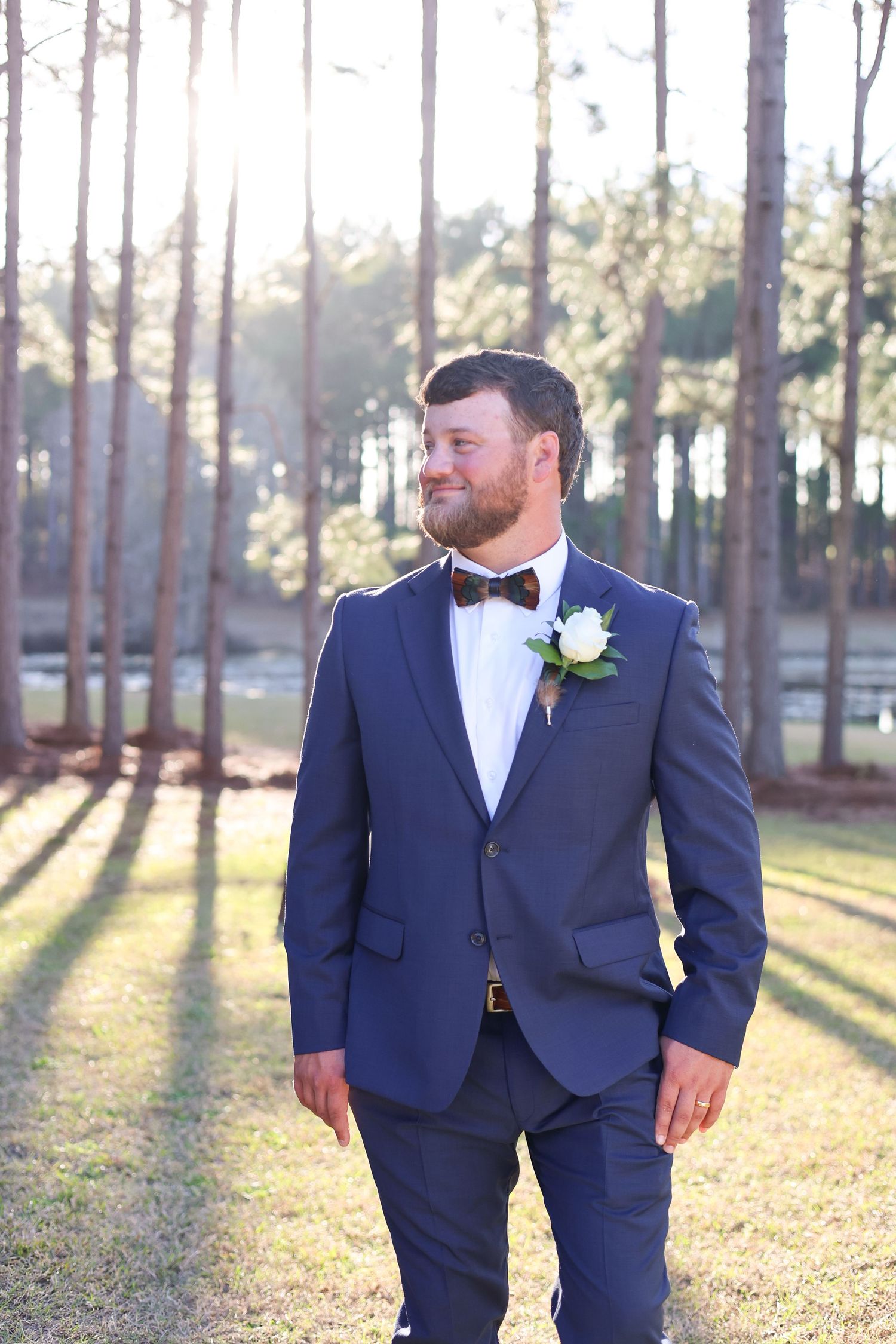 A groom in a navy blue suit with bowtie and boutonniere poses outdoors among sunlit pine trees.
