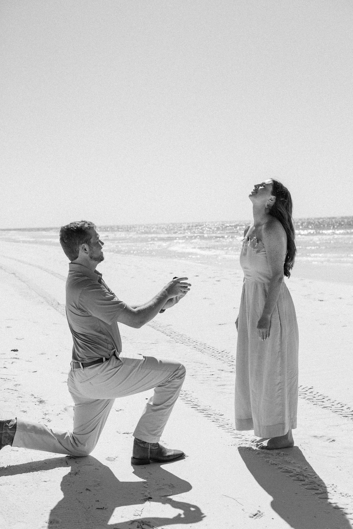 Black and white photo of a man on one knee proposing to a woman on a sunny beach.