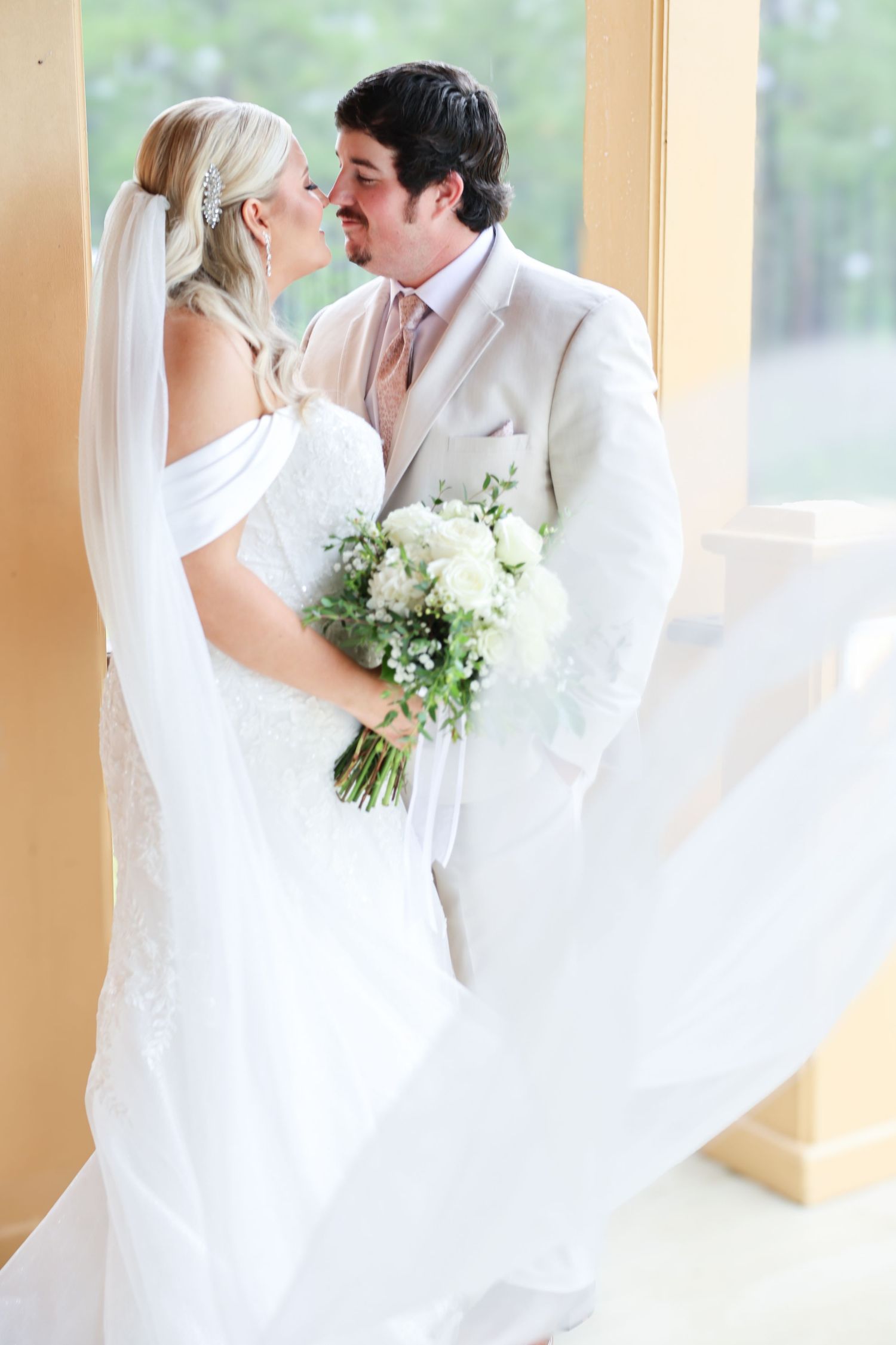 Bride and groom nearly kissing with flowing veil and white floral bouquet in romantic indoor setting.