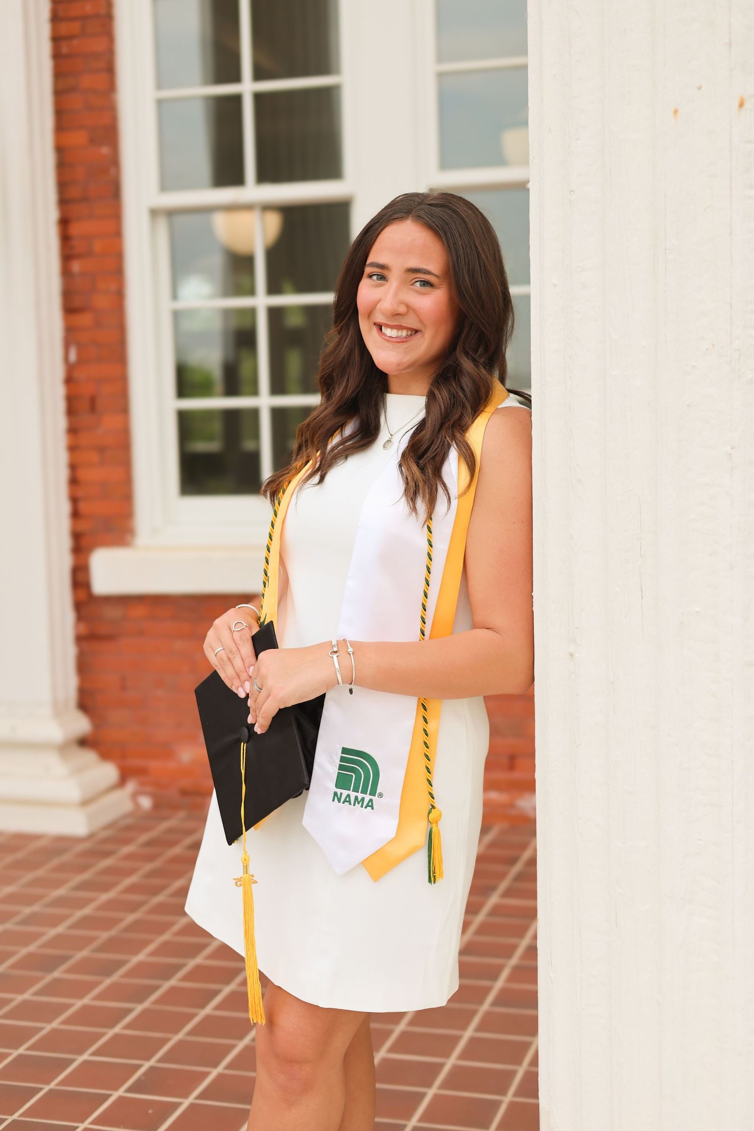 Graduate in white dress with gold honors stole poses between white columns.