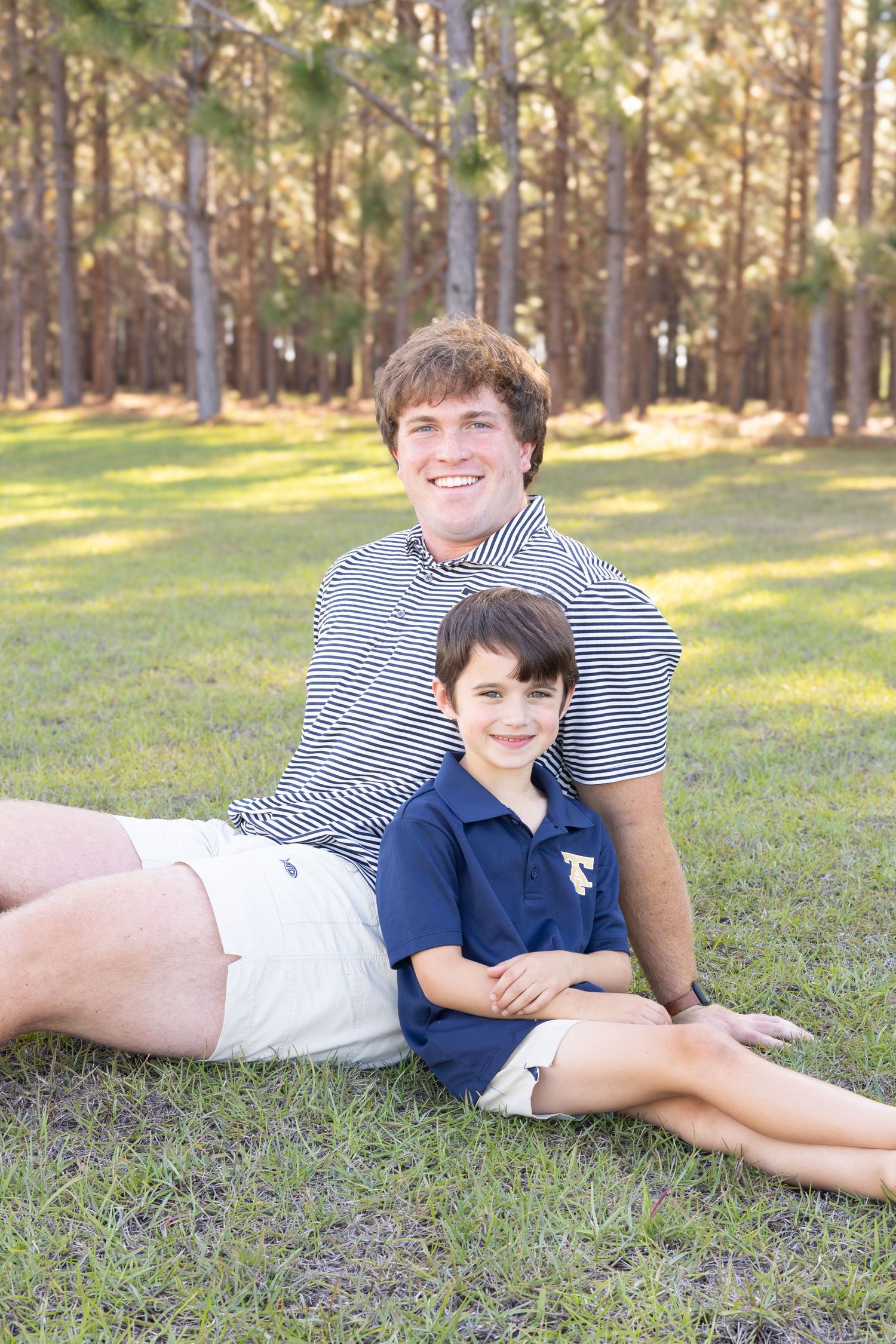 Two boys smiling and relaxing on green grass in front of tall pine trees, wearing navy and striped polo shirts.
