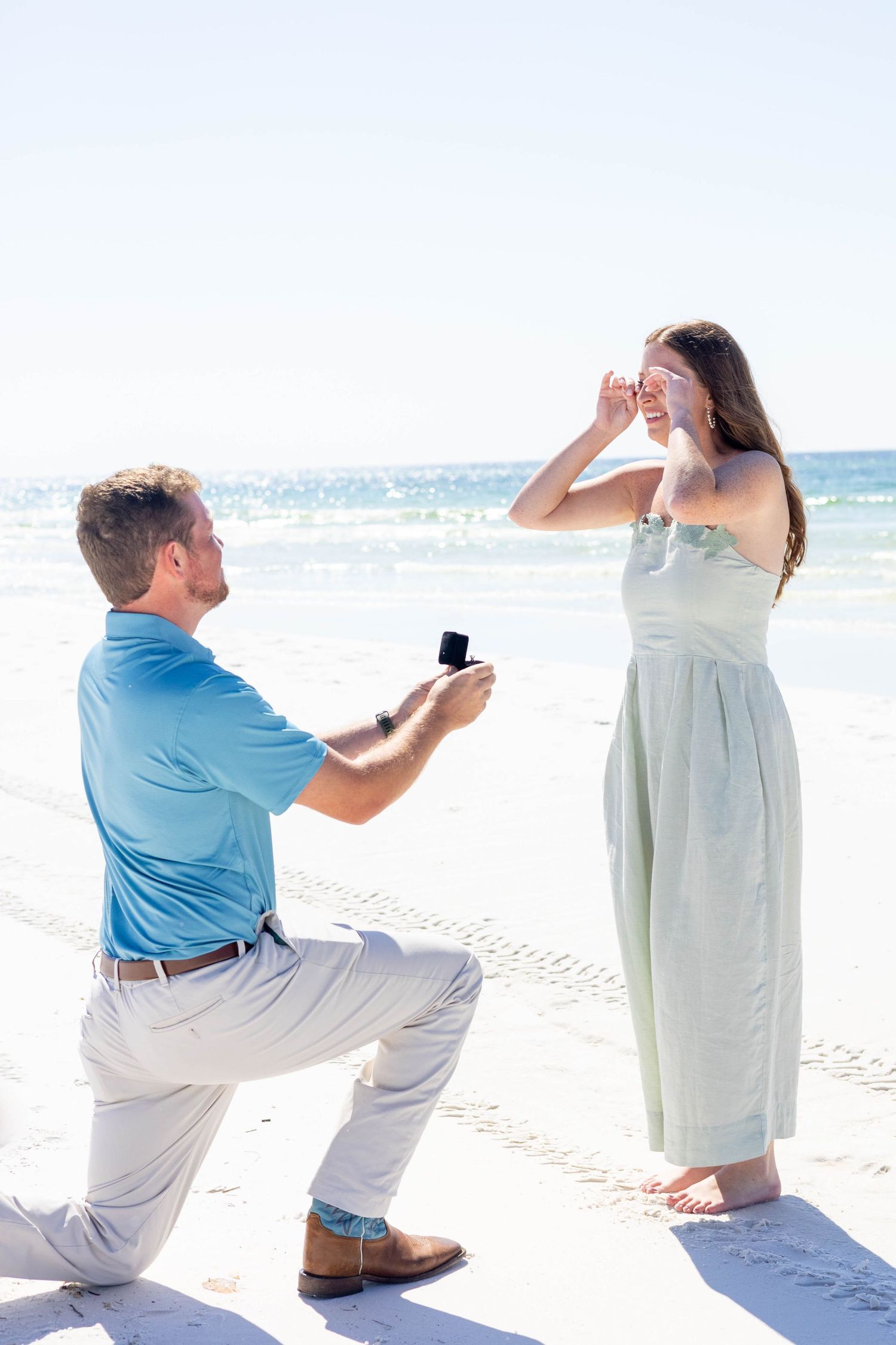 Man kneeling on beach proposing with ring box to surprised woman in green dress by the ocean.