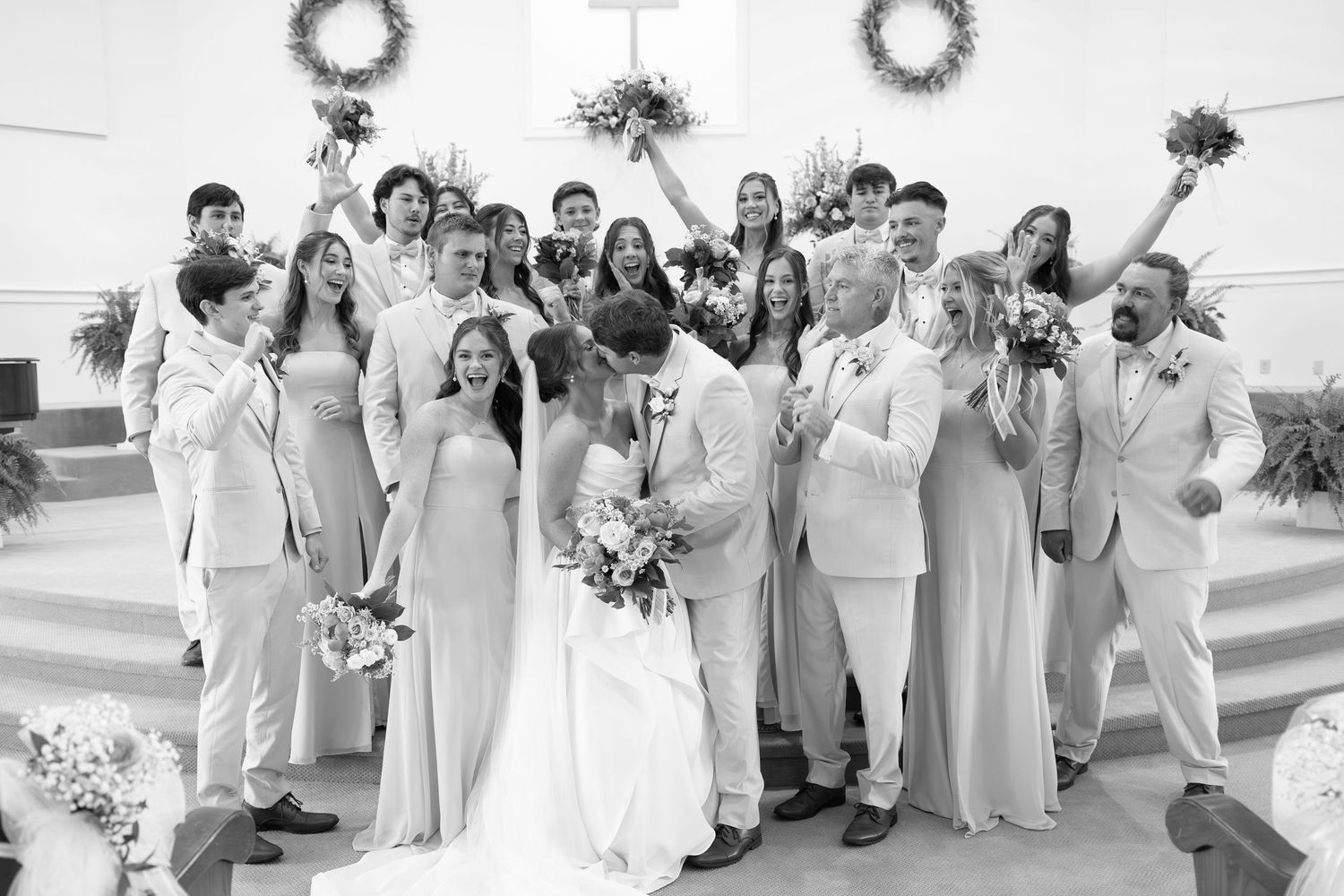 Black and white wedding photo of bride and groom kissing surrounded by smiling bridal party in church.