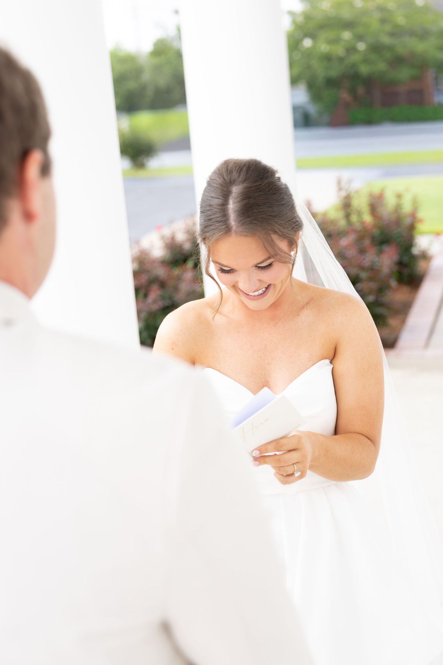 Smiling bride in strapless wedding gown and veil reading vows from a booklet during outdoor ceremony.