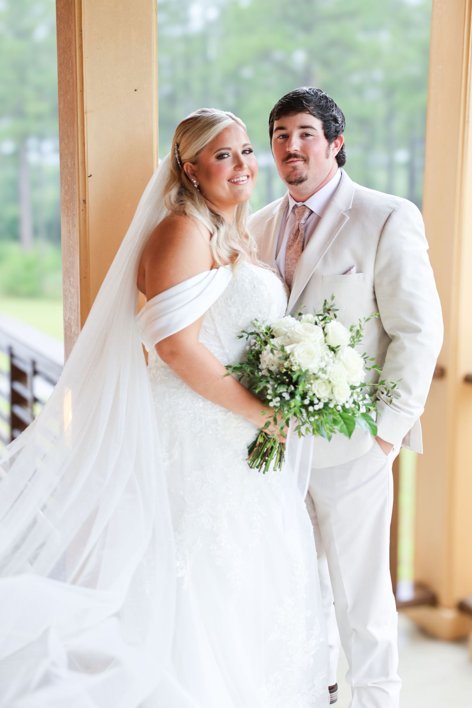 Bride and groom pose together indoors, she in lace off-shoulder gown with veil, he in cream suit with pink tie.
