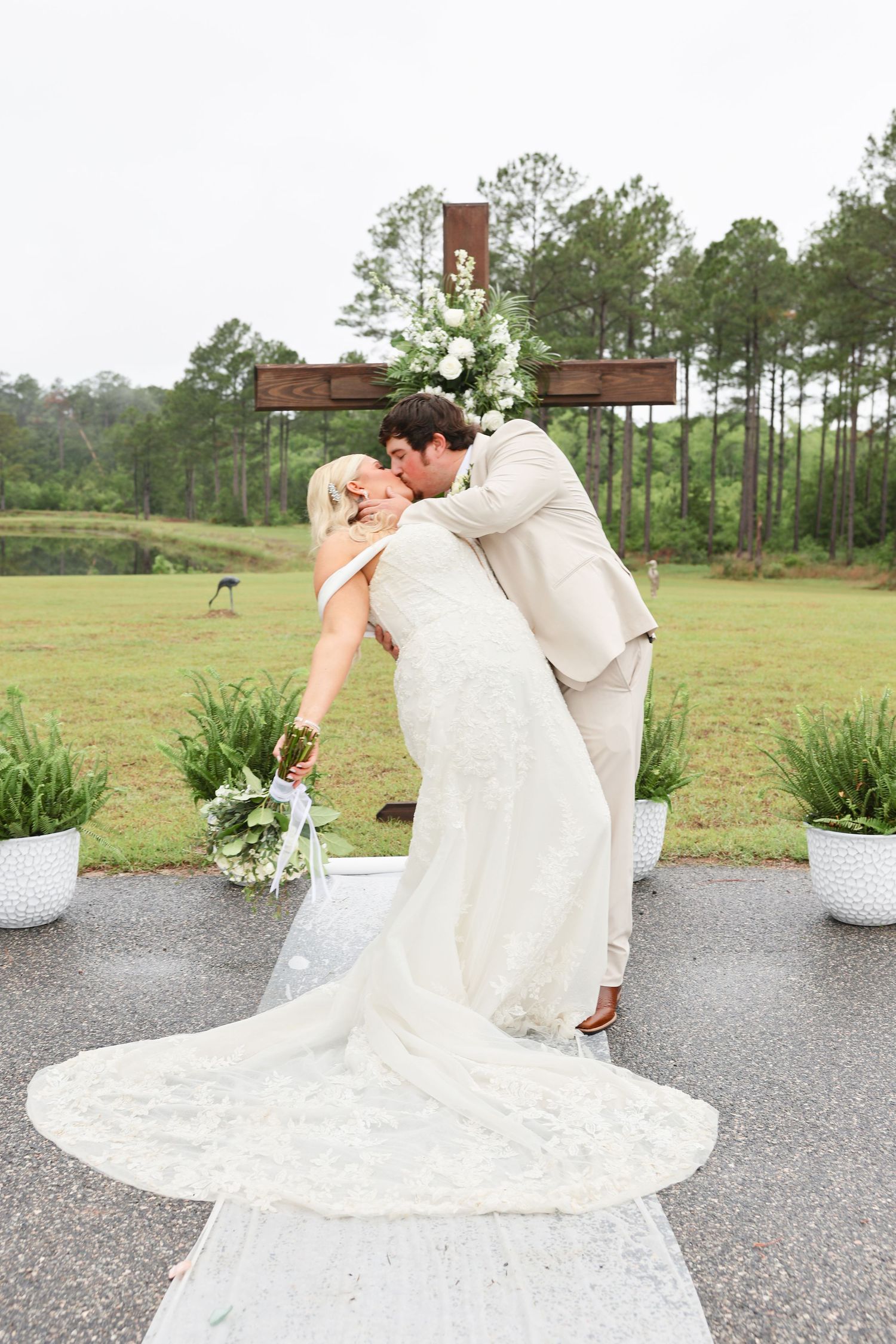 Groom dips bride for a romantic kiss at outdoor wedding ceremony with wooden cross and white floral arrangements.