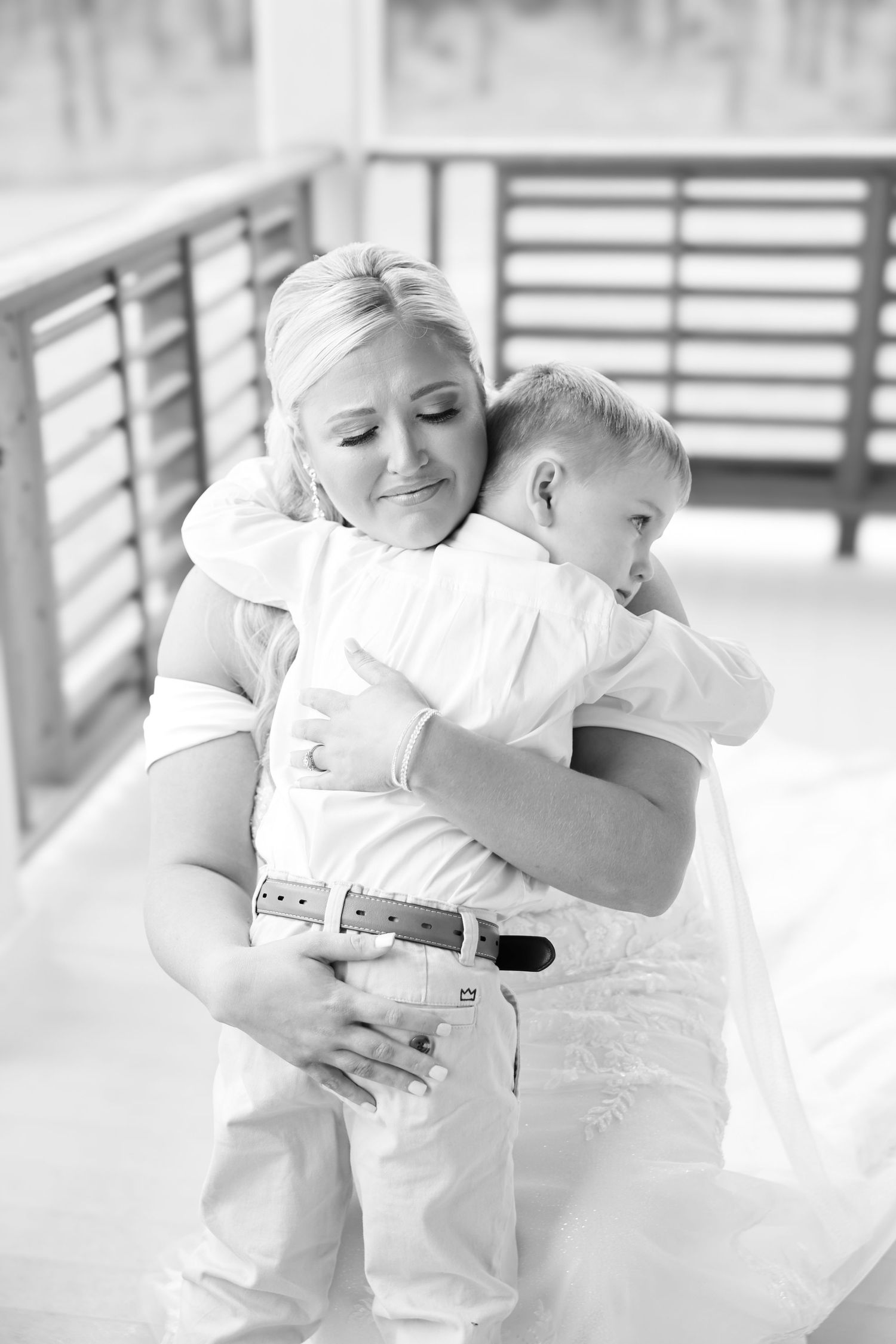 Bride in wedding dress sharing tender hug with young boy in white outfit on outdoor deck.