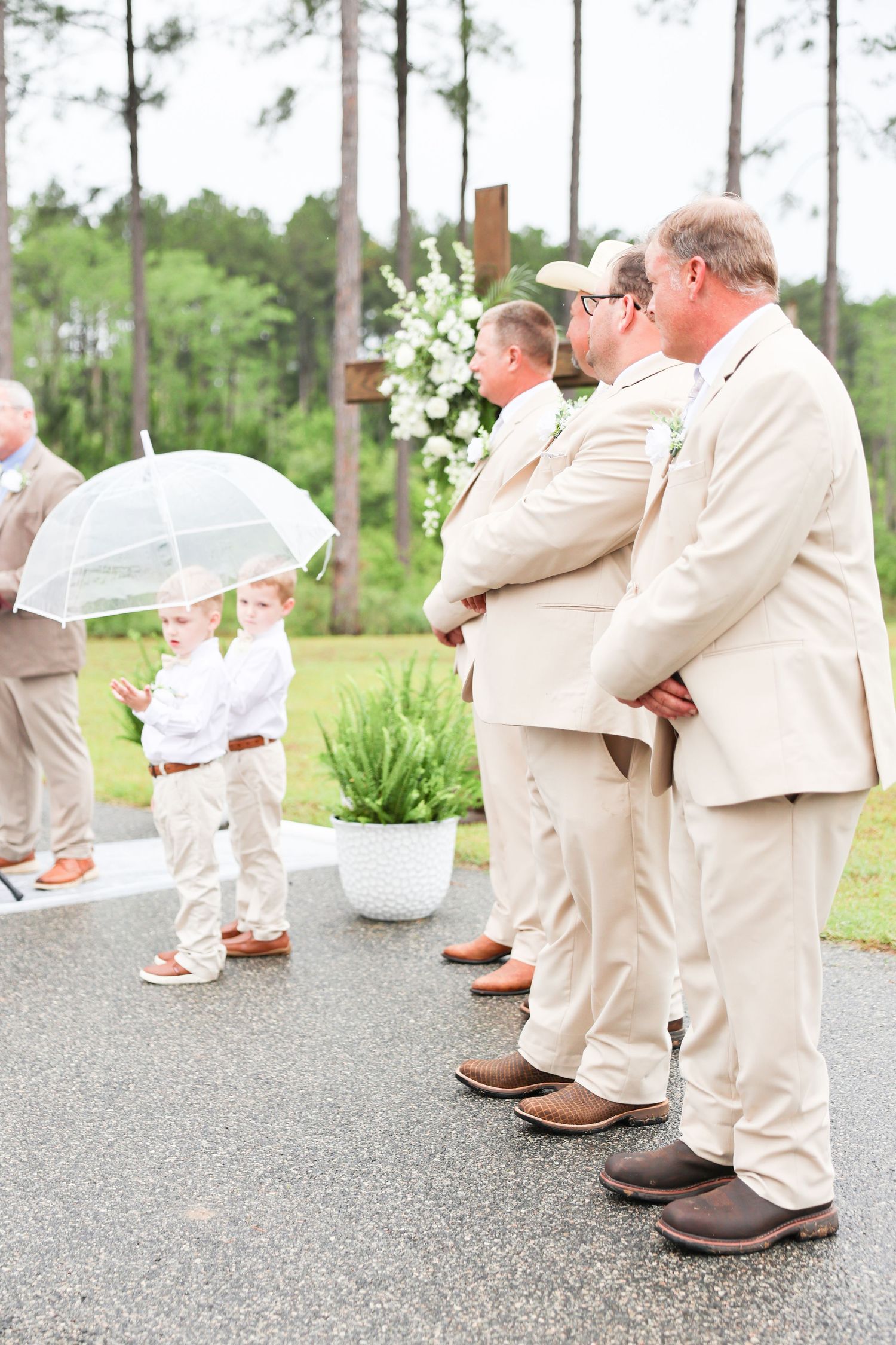 Young ring bearer with clear umbrella stands near groomsmen in tan suits at outdoor wedding ceremony.